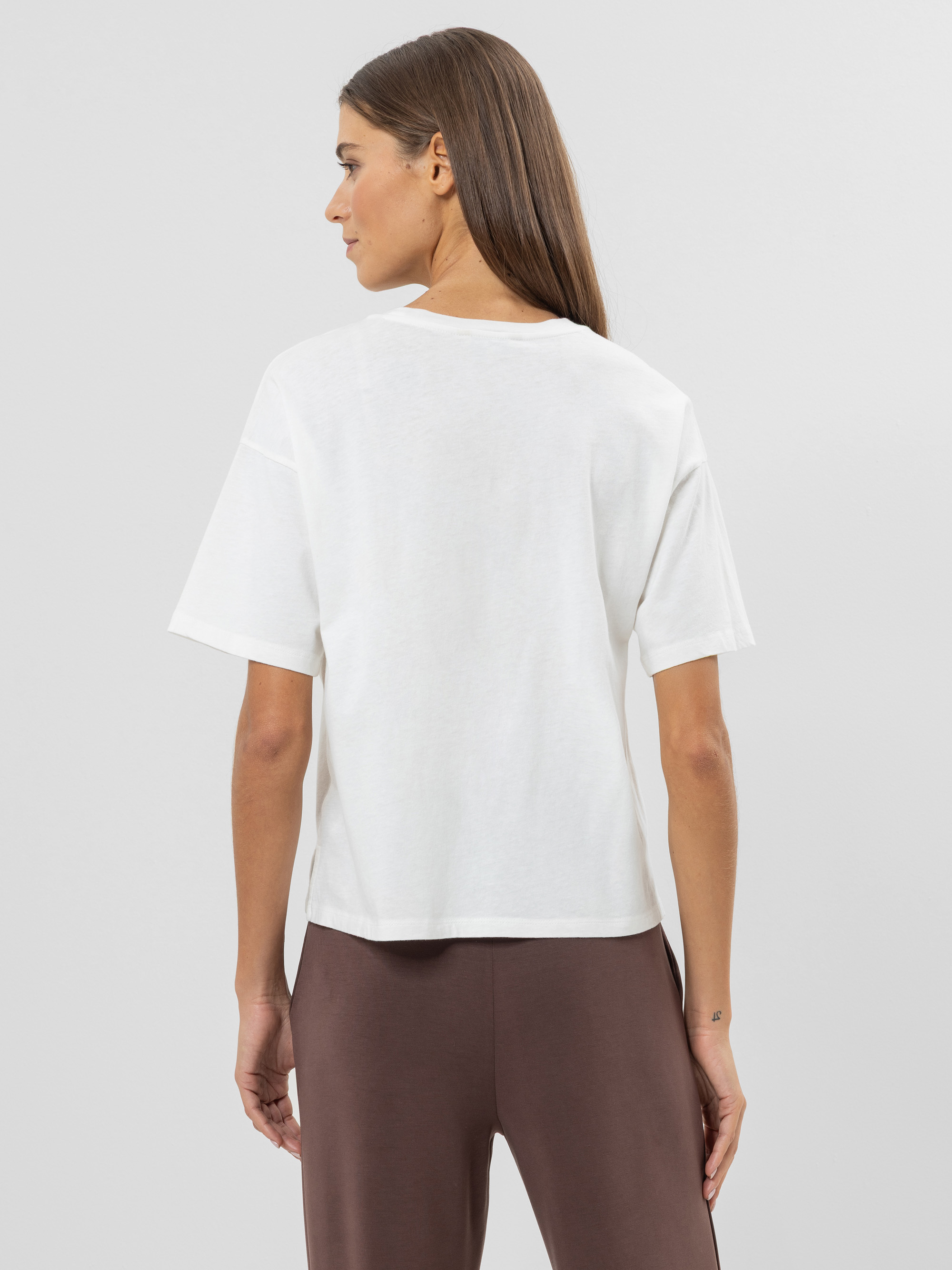 Eine Frau mit glattem braunem Haar steht mit dem Gesicht zur Wand, trägt ein lockeres weißes T-Shirt und eine braune Hose vor einem schlichten hellgrauen Hintergrund.