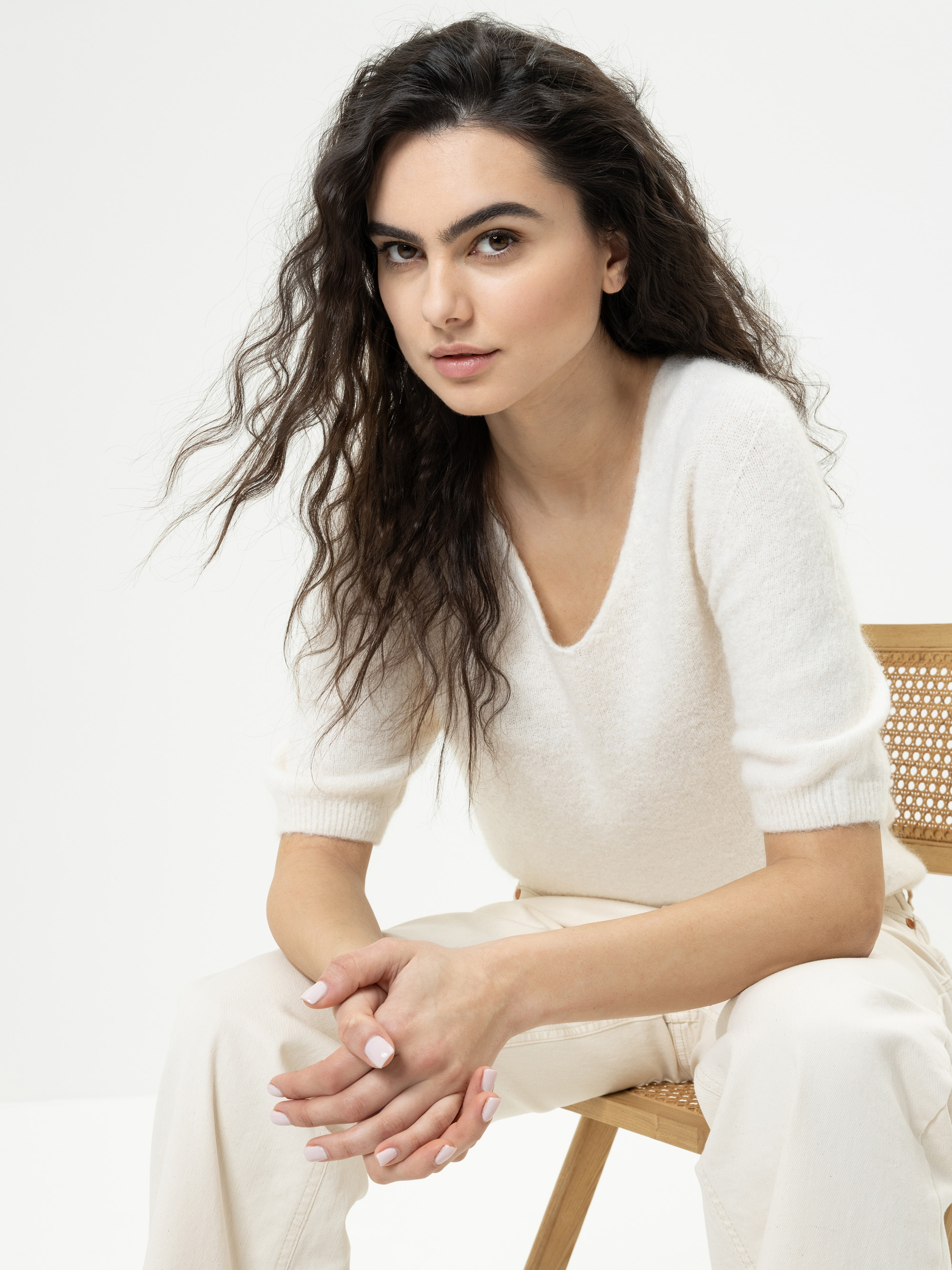 Eine Frau mit langen, gewellten dunklen Haaren, die ein weißes Strickoberteil und eine helle Hose trägt, sitzt auf einem Holzstuhl und blickt mit einem leichten Lächeln in die Kamera vor einem schlichten weißen Hintergrund.