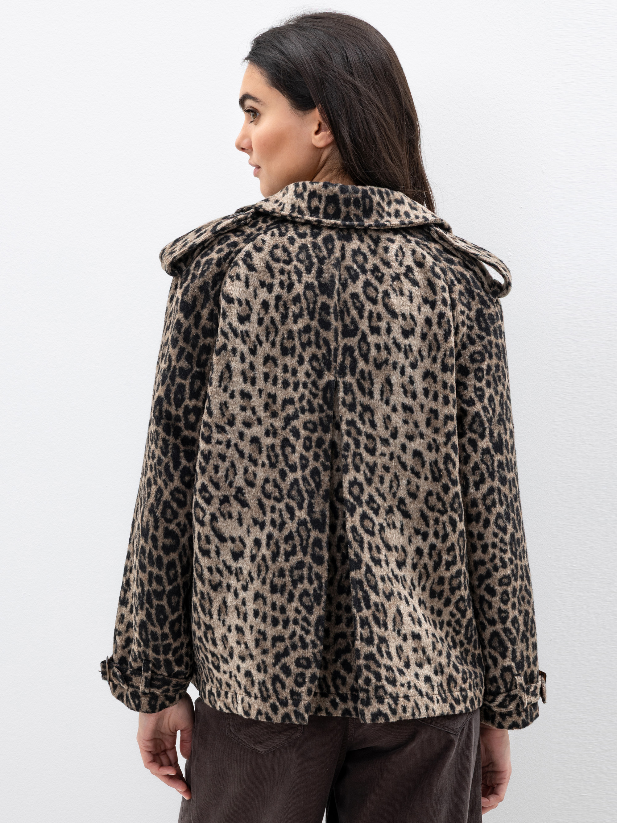Eine Frau steht mit dem Gesicht zur Wand und trägt einen braun-schwarzen Mantel mit Leopardenmuster, Kragen und Knopfleiste. Ihr dunkles Haar ist offen, und sie trägt eine dunkle Hose vor einem schlichten weißen Hintergrund.