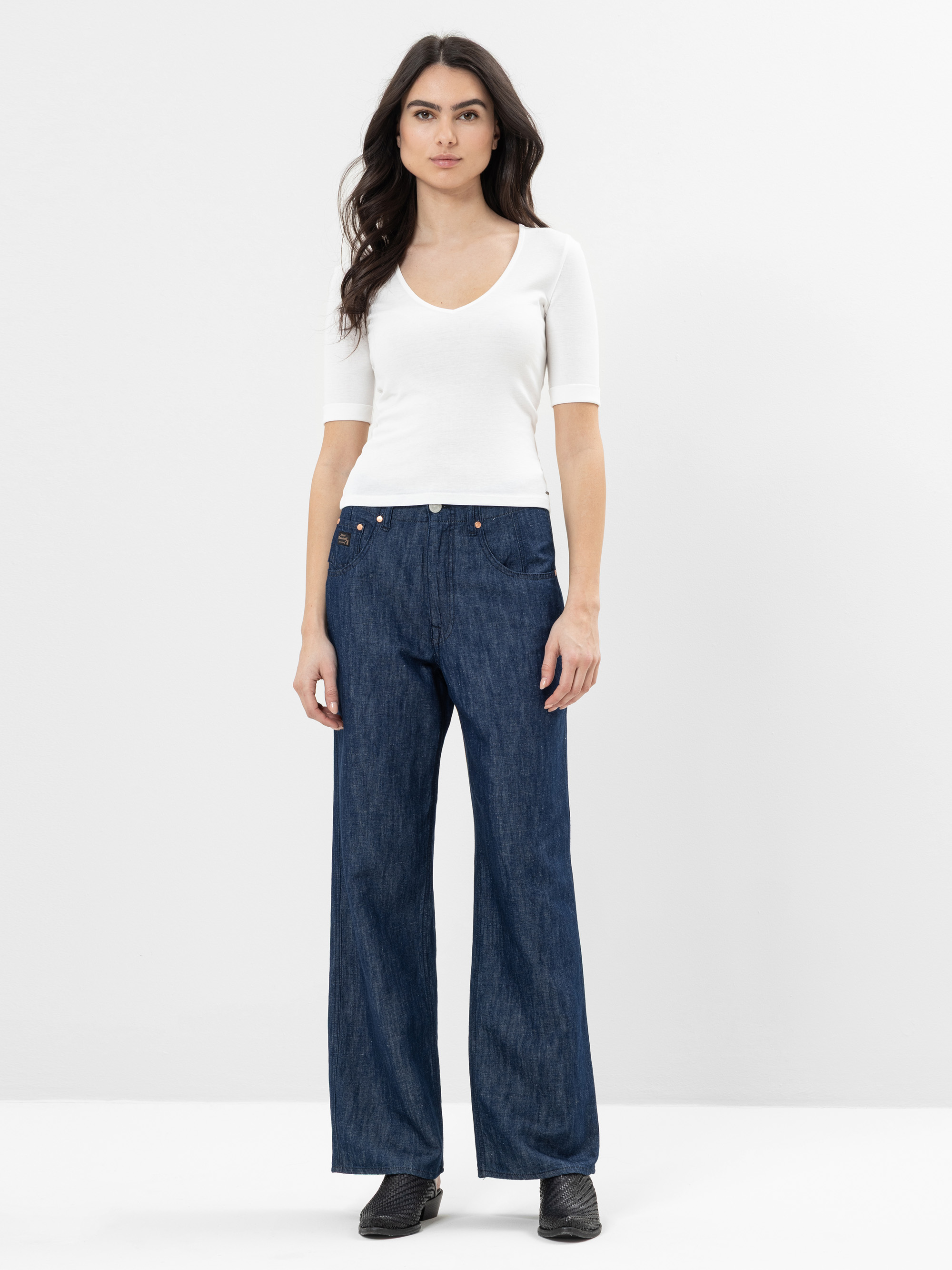 Eine Frau steht vor einem schlichten weißen Hintergrund. Sie trägt ein kurzärmeliges weißes Oberteil, weite Jeans und schwarze Schuhe, ihr langes dunkles Haar fällt nach unten und sie hat die Hände an den Seiten.