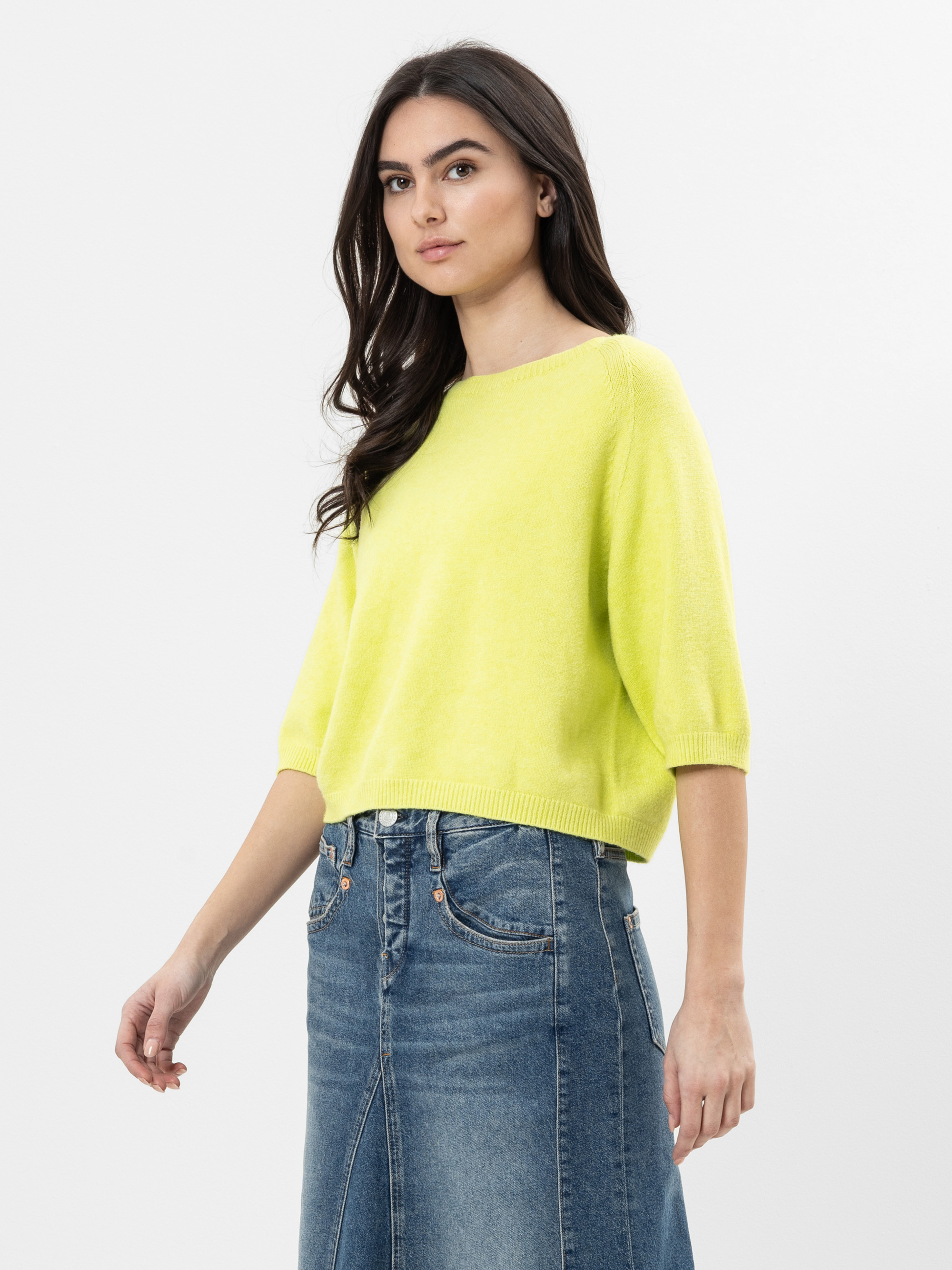 Eine Frau mit langen dunklen Haaren trägt einen leuchtend gelben kurzärmeligen Pullover und einen blauen Jeansrock und steht vor einem schlichten weißen Hintergrund.