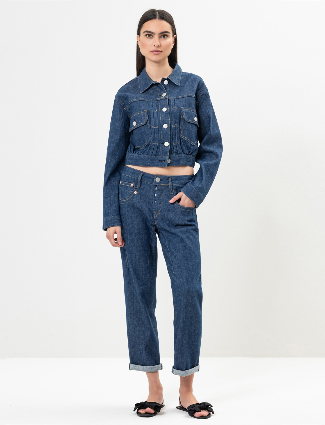 Eine Frau steht vor einem schlichten weißen Hintergrund und trägt eine abgeschnittene blaue Jeansjacke und passende blaue Jeans mit aufgerollten Bündchen. Sie hat langes dunkles Haar und ist barfuß, abgesehen von schwarzen Schleifen-Sandalen.