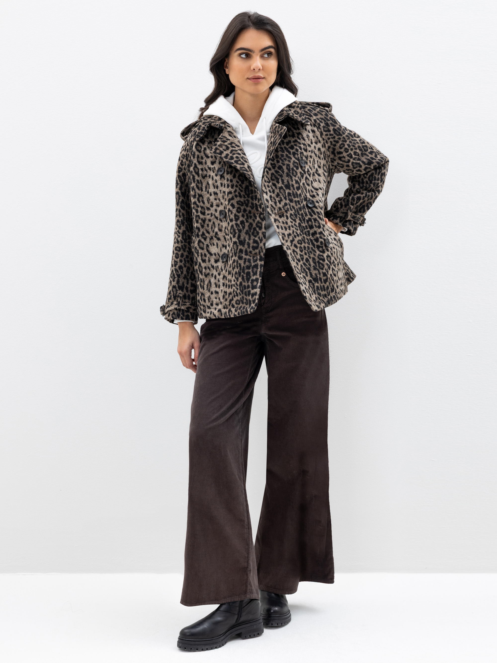 Eine Frau steht vor einem weißen Hintergrund und trägt eine Jacke mit Leopardenmuster, ein weißes Hemd, eine braune Hose mit weitem Bein und schwarze Stiefel. Sie hat langes dunkles Haar und eine Hand in die Hüfte gestemmt und posiert selbstbewusst.