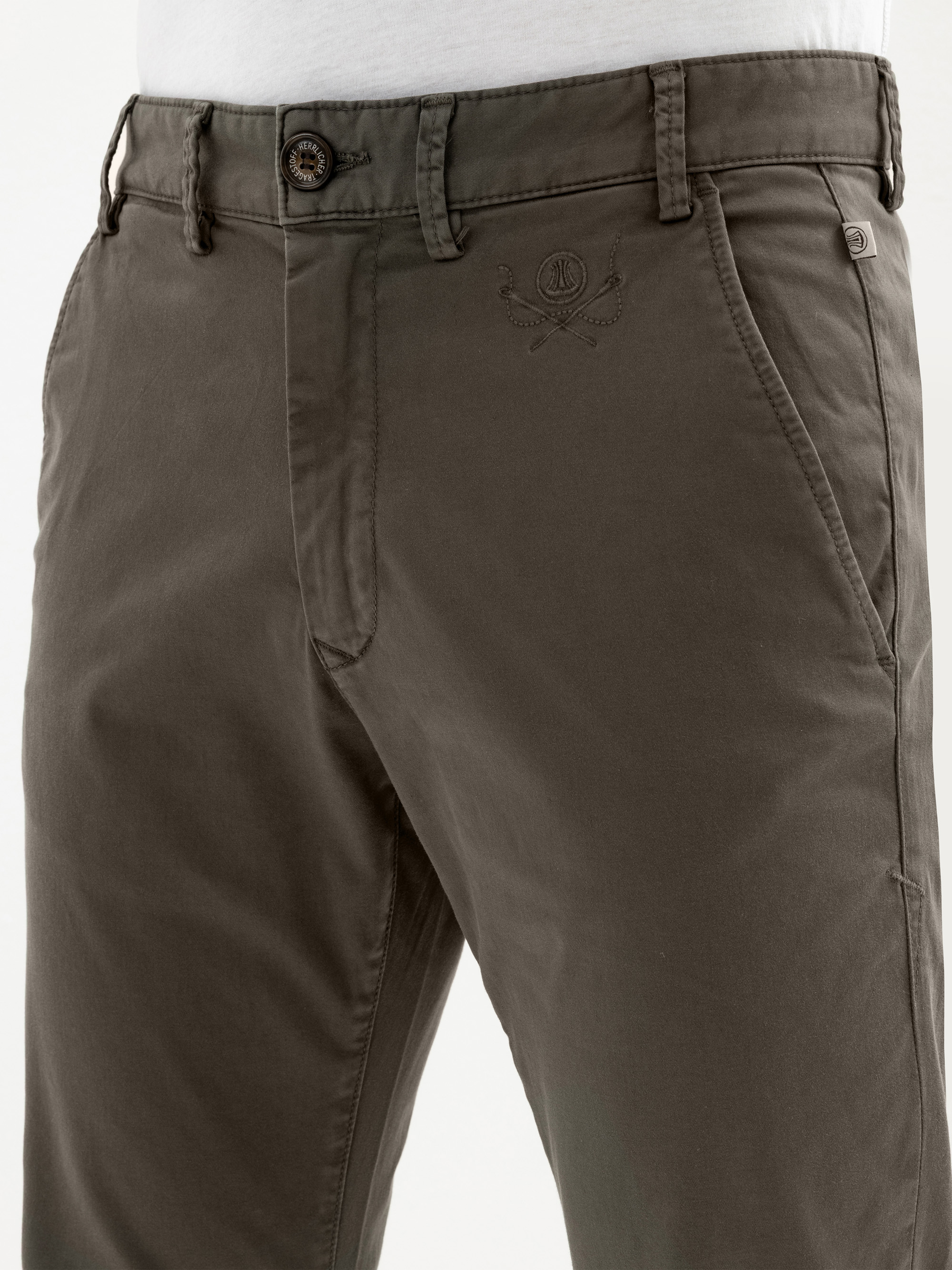 Nahaufnahme einer Person, die eine olivgrüne Hose mit Knopf und Reißverschluss, schrägen Vordertaschen und einem gestickten Logo auf der rechten Seite unterhalb des Hosenbundes trägt. Ein weißes Hemd ist teilweise über der Hose sichtbar.