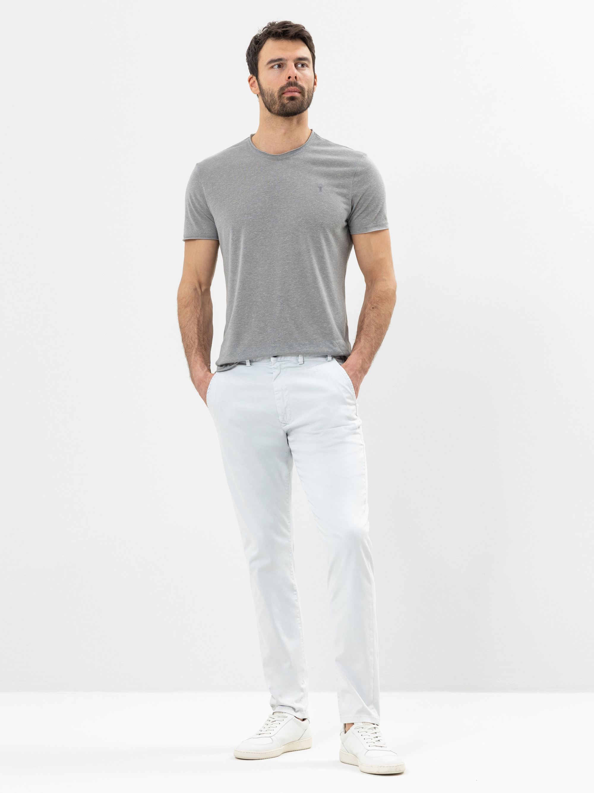 Ein Mann steht vor einem schlichten weißen Hintergrund, trägt ein graues T-Shirt, eine helle Hose und weiße Turnschuhe und hat die Hände in den Taschen.