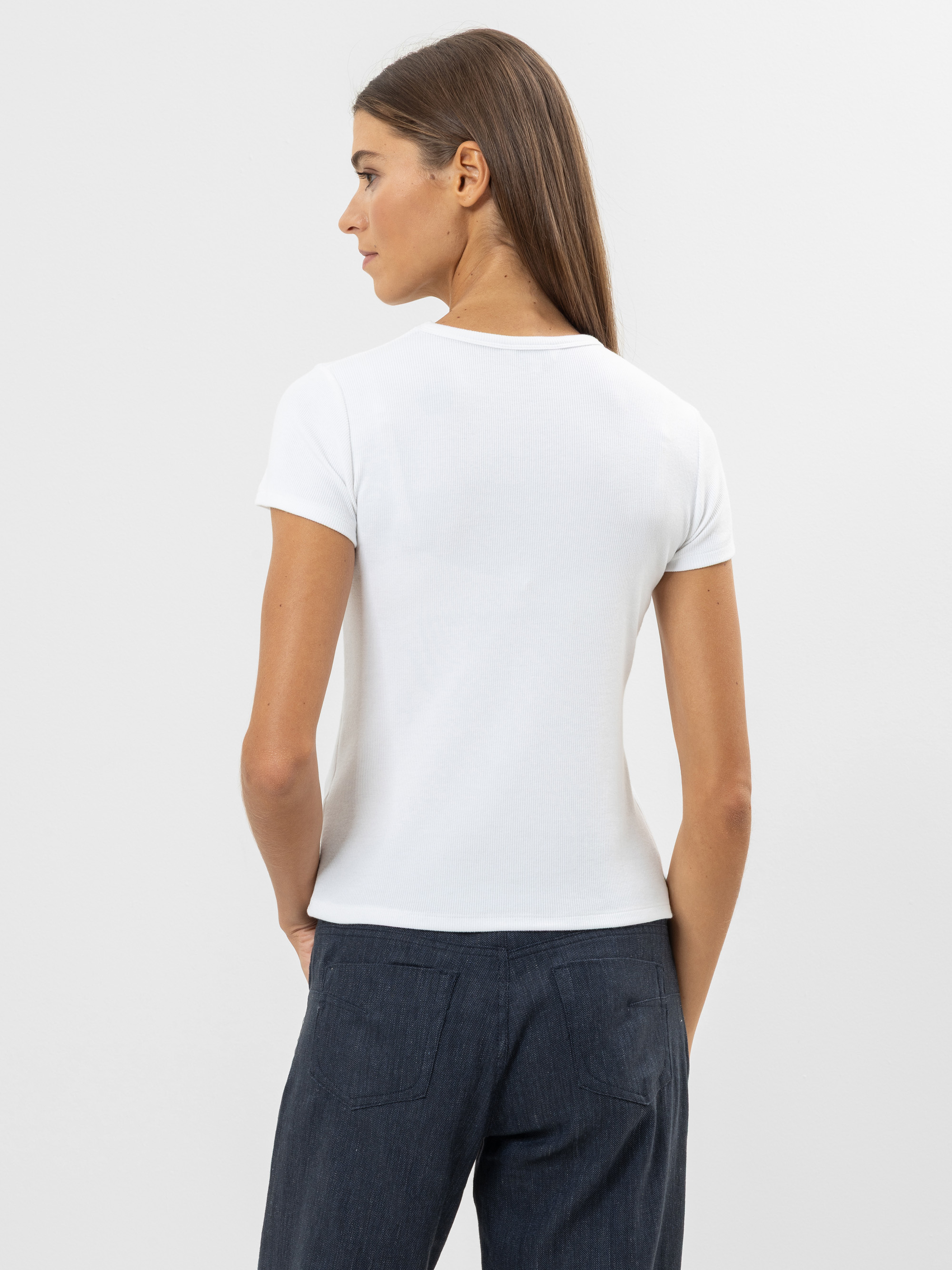 Eine Frau mit langen braunen Haaren steht mit dem Gesicht zur Wand und trägt ein weißes, kurzärmeliges T-Shirt und eine dunkle Hose vor einem schlichten, hellen Hintergrund.