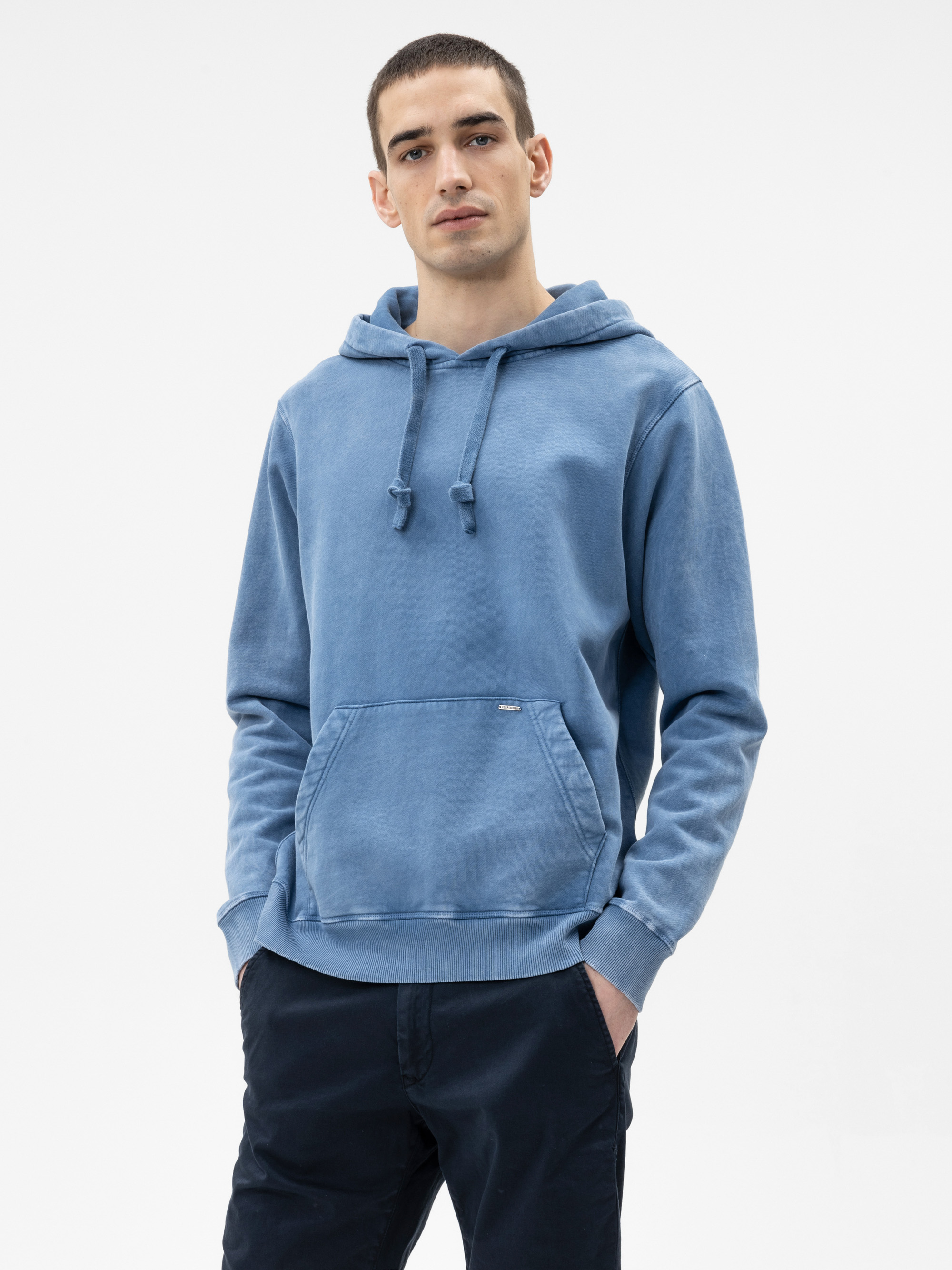 Eine Person steht vor einem schlichten weißen Hintergrund, trägt einen hellblauen Kapuzenpulli mit einer Vordertasche und eine dunkle Hose und blickt mit den Händen in den Taschen direkt in die Kamera.