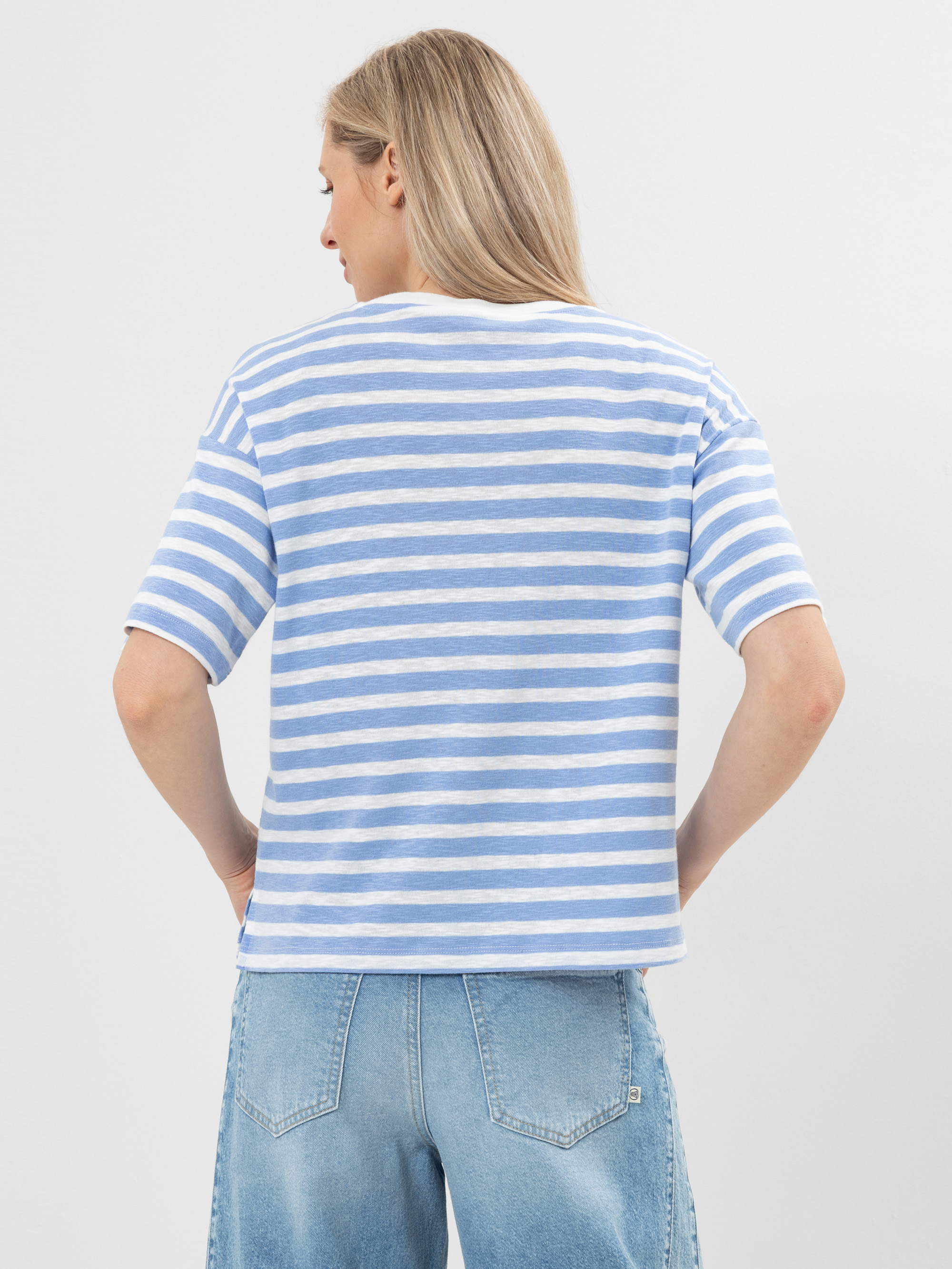 Eine Person mit langen blonden Haaren, von hinten gesehen, trägt ein hellblau-weiß gestreiftes kurzärmeliges Hemd und hellblaue Jeans. Ihre Hände ruhen auf den Hüften vor einem schlichten hellen Hintergrund.