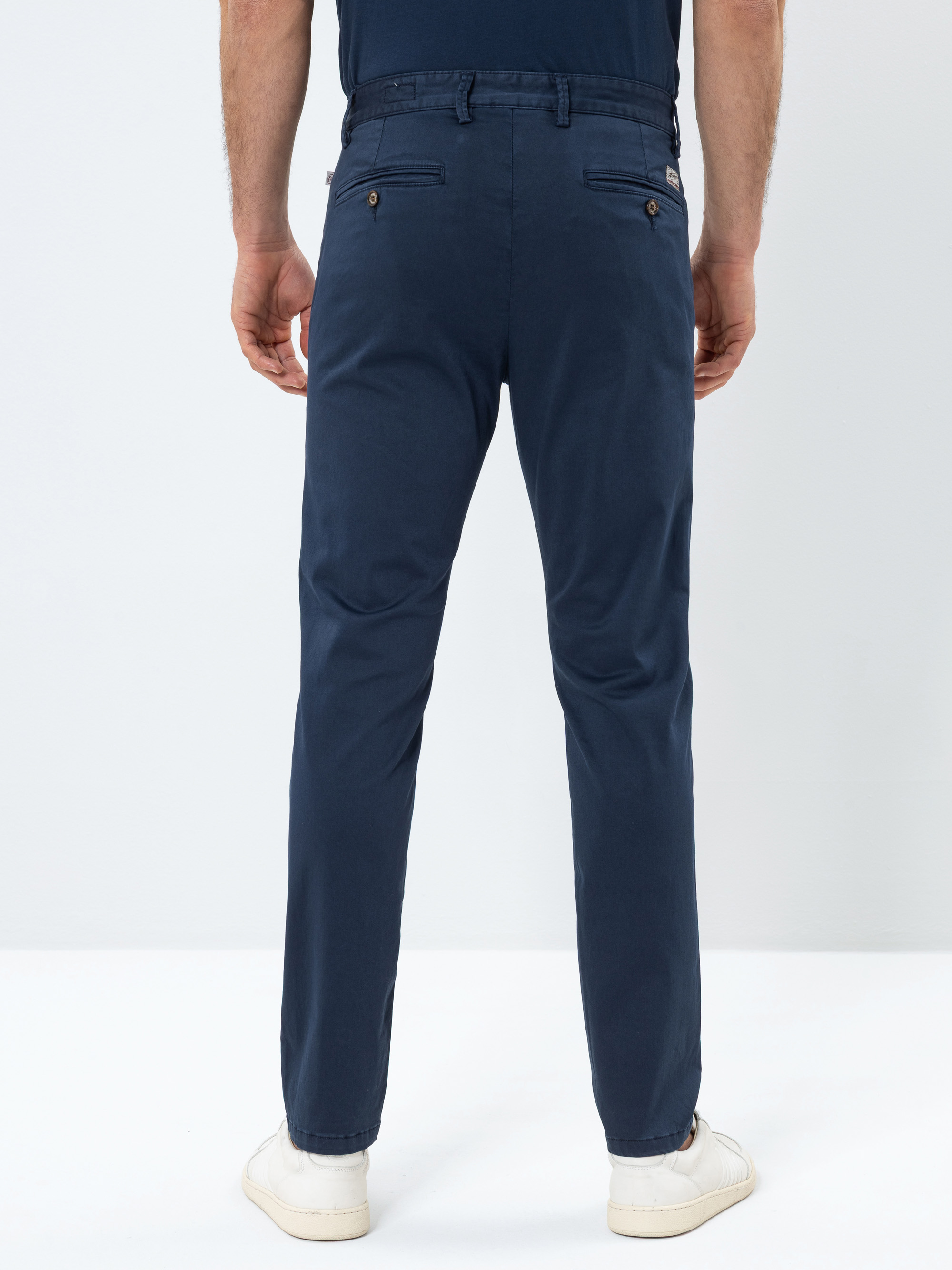 Ein Mann steht mit dem Rücken zur Kamera, trägt eine marineblaue Hose, einen passenden Gürtel und weiße Turnschuhe vor einem schlichten weißen Hintergrund.