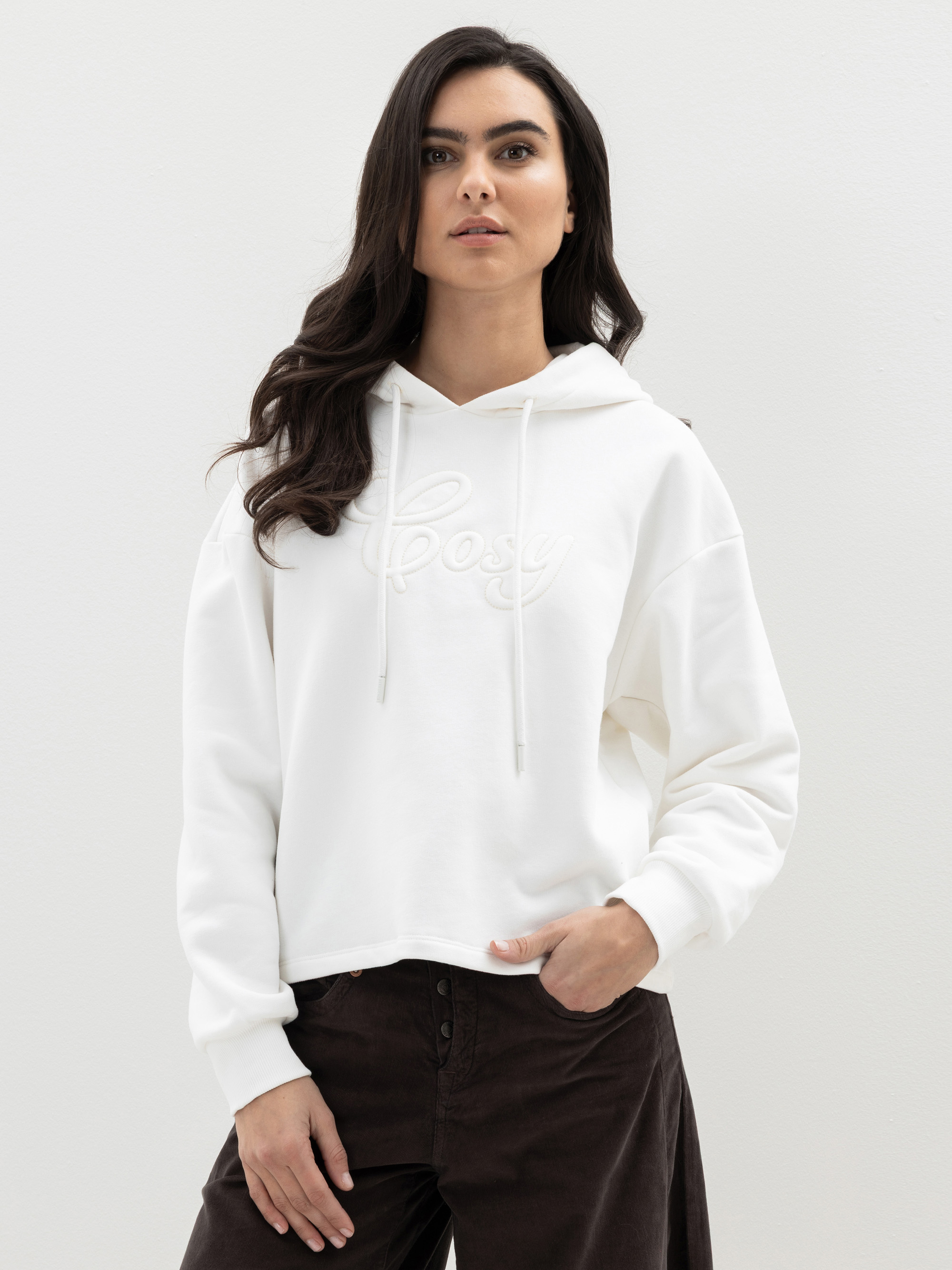 Eine Frau mit langen dunklen Haaren trägt einen weißen Kapuzenpulli mit dem aufgestickten Wort Cosy und eine dunkle Hose und steht vor einem schlichten hellen Hintergrund.