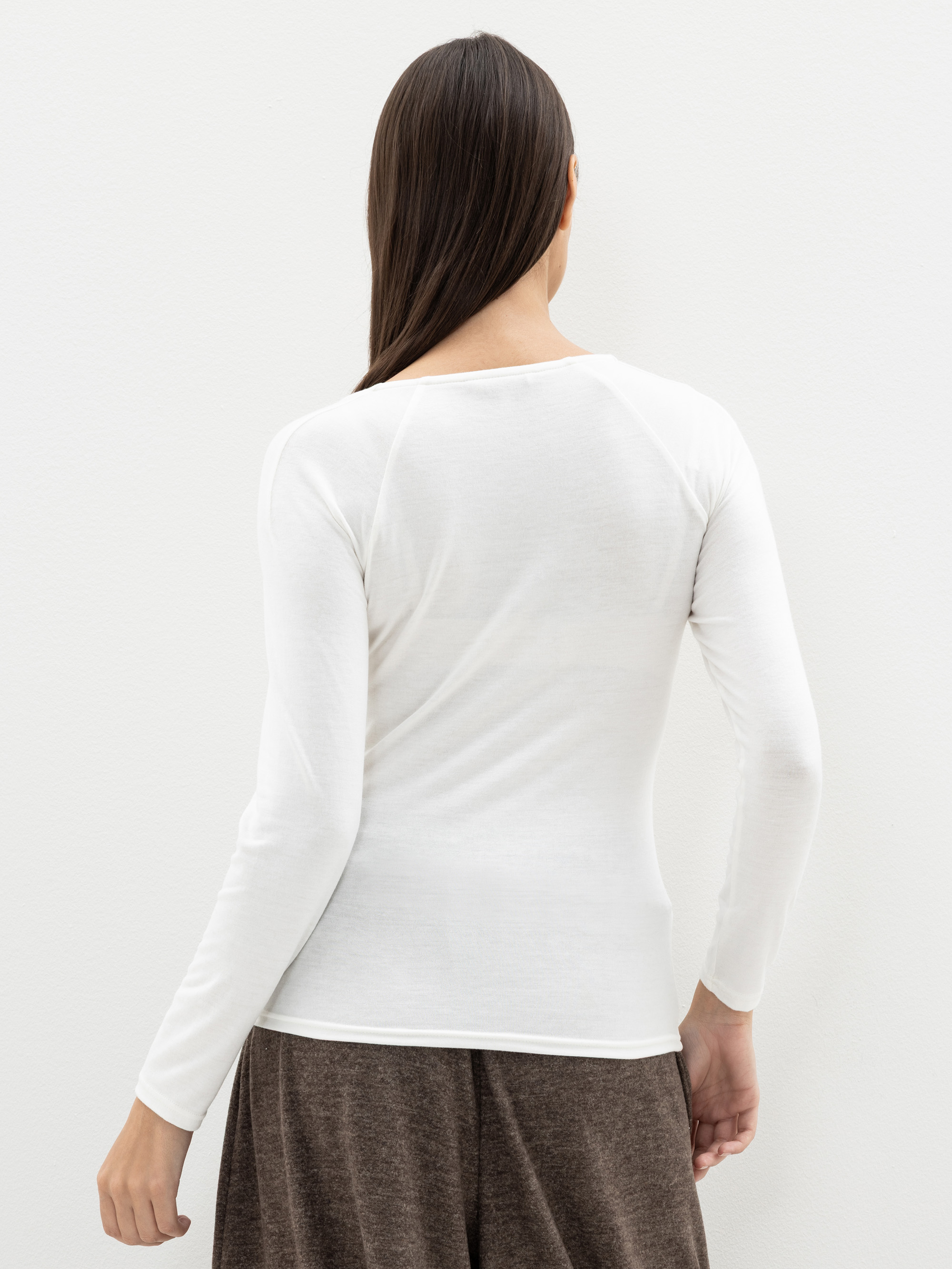 Eine Person mit langen braunen Haaren, von hinten gesehen, trägt ein weißes, langärmeliges Hemd und eine weite braune Hose und steht vor einem schlichten weißen Hintergrund.