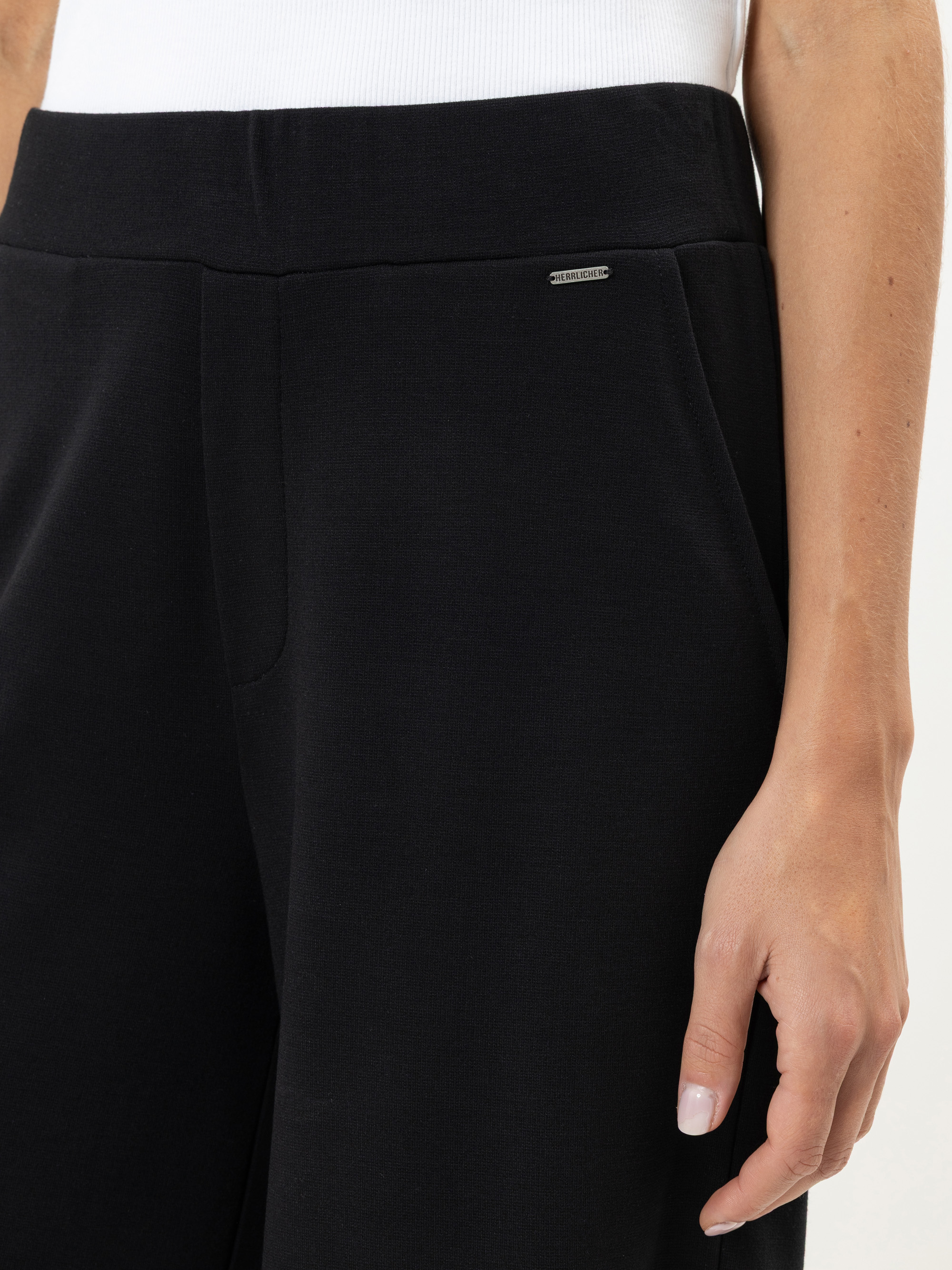 Nahaufnahme einer Person, die eine schwarze Hose und ein weißes Oberteil trägt und eine Hand an der Seite ruhen lässt. Die Hose hat ein kleines Metalllogo in der Nähe des Hosenbundes.