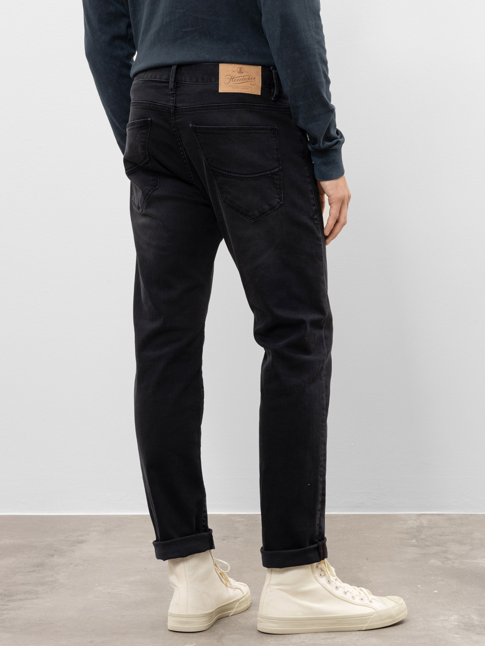 Eine Person, die eine schwarze Jeans, ein dunkles langärmeliges Hemd und weiße High-Top-Turnschuhe trägt, steht mit dem Gesicht zur Seite und zeigt die Rückseite ihrer Jeans mit einem braunen Fleck am Bund.
