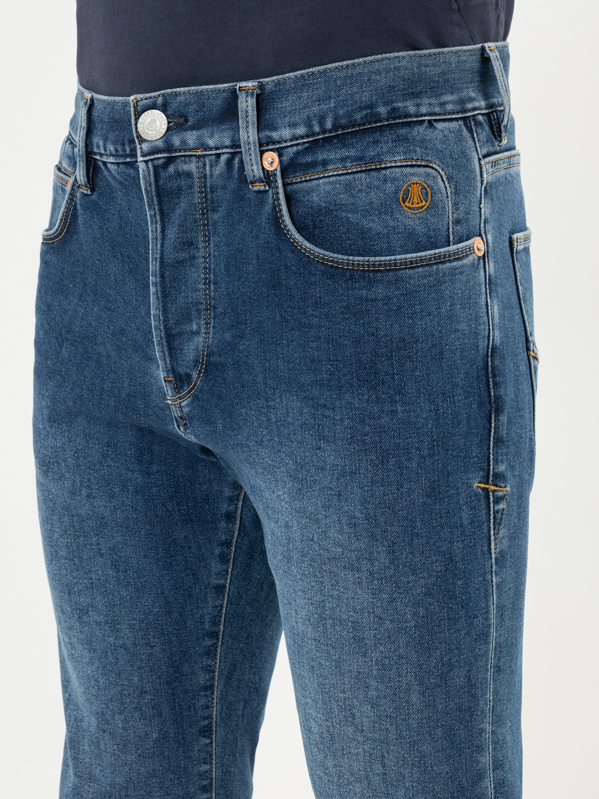 Eine Person, die eine blaue Jeans mit orangefarbenen Nähten und einem kleinen runden Logo auf der Vordertasche trägt, gepaart mit einem dunklen Hemd.