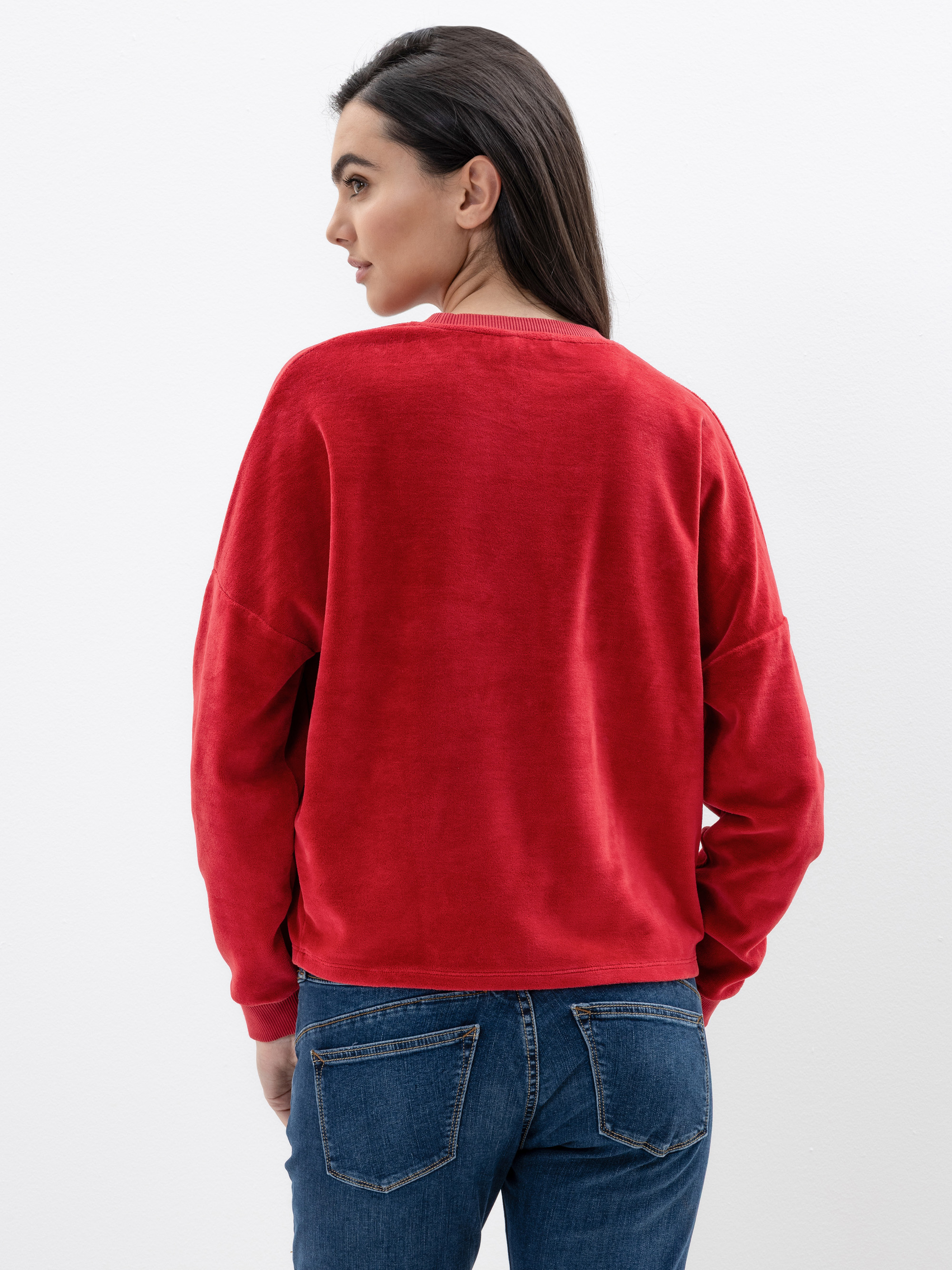 Eine Frau mit langen dunklen Haaren, die einen roten Pullover und blaue Jeans trägt, steht mit dem Gesicht zur Kamera vor einem schlichten weißen Hintergrund.