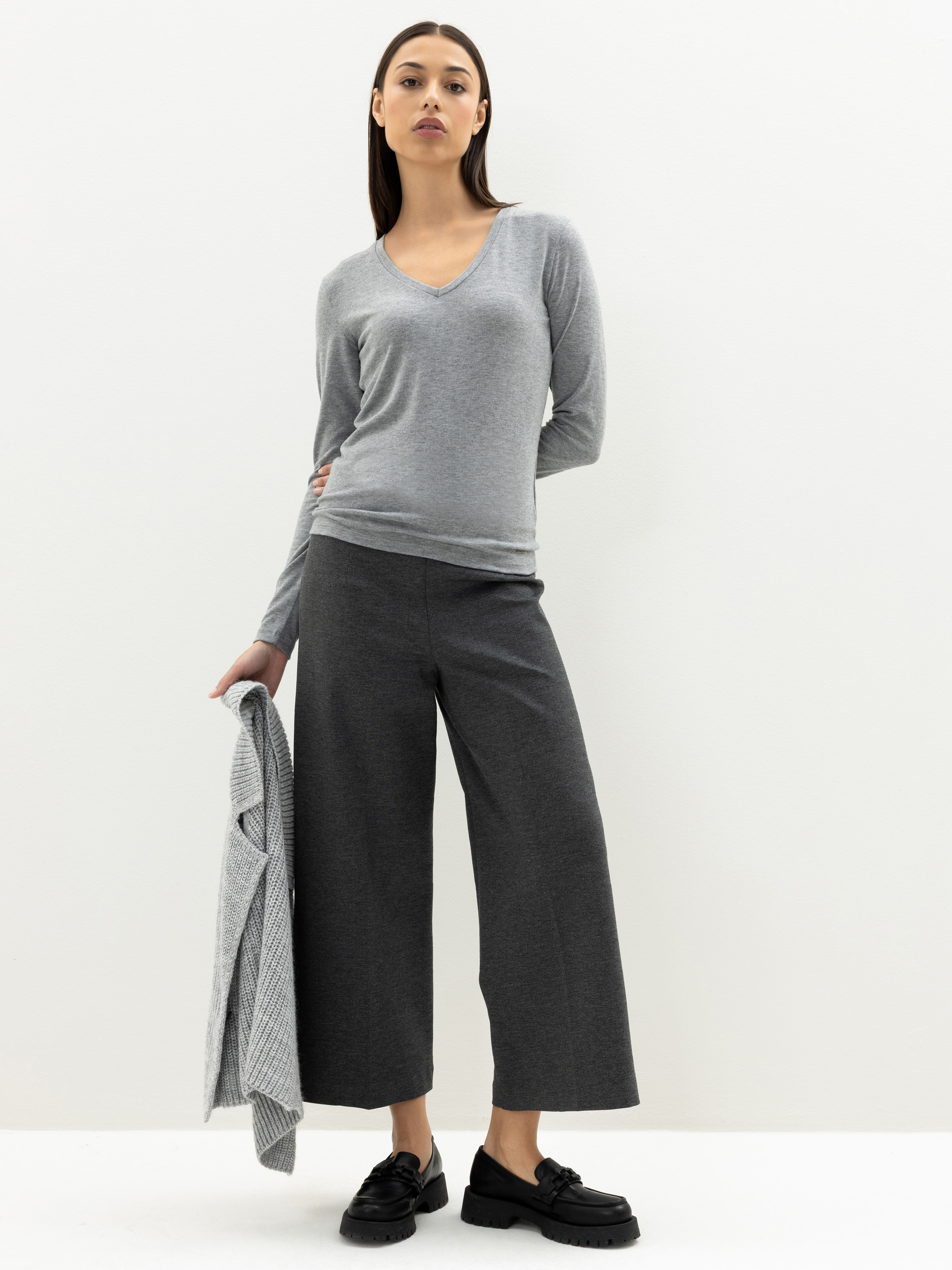 Eine Frau steht vor einem weißen Hintergrund und trägt einen hellgrauen Pullover mit V-Ausschnitt, eine dunkelgraue Hose mit weitem Bein und schwarze Schuhe. In ihrer linken Hand hält sie eine grau karierte Jacke.