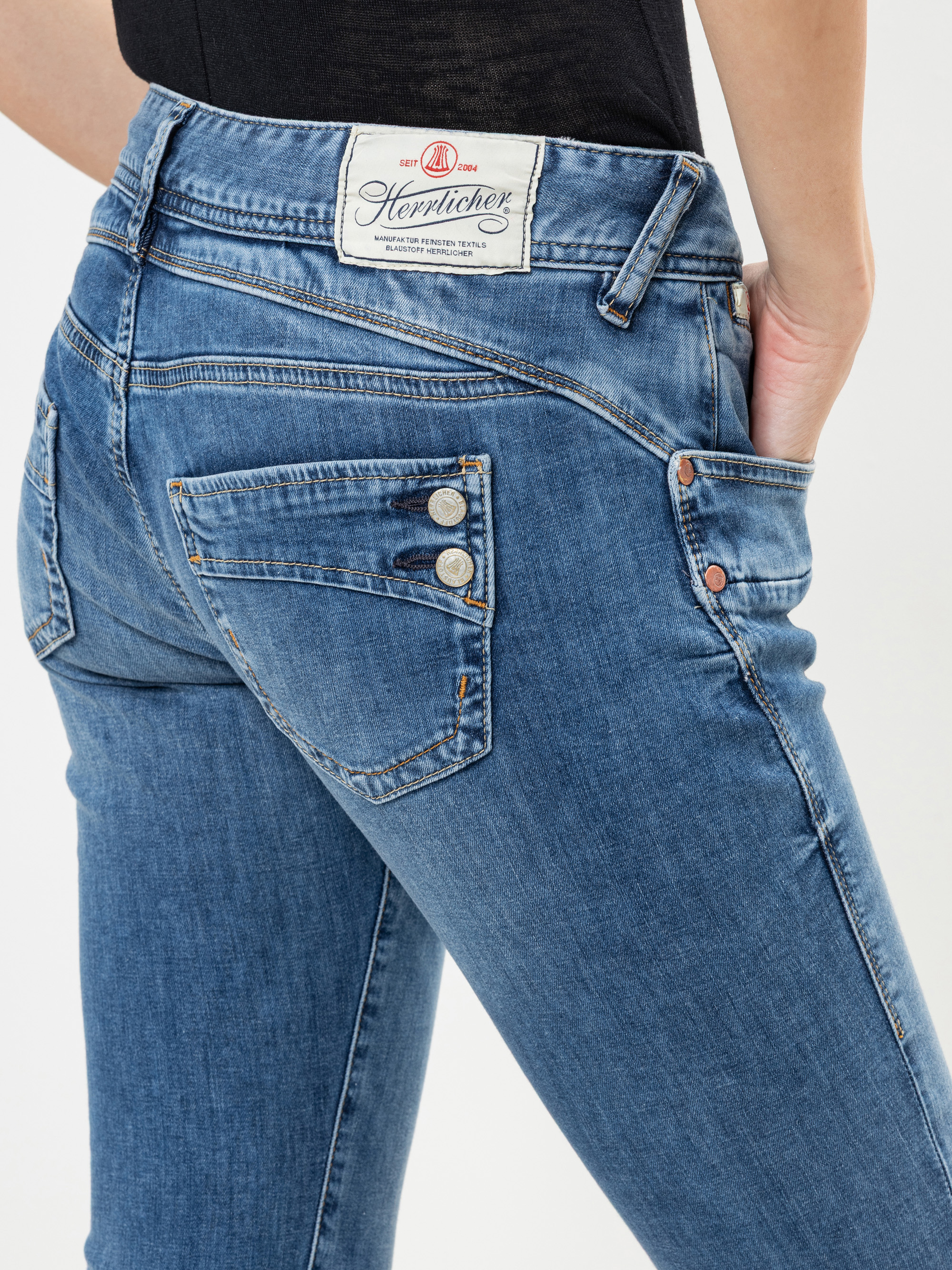 Person, die eine blaue Denim-Jeans mit einem weißen Herrlicher-Markenetikett am Bund, einer dekorativen Gesäßtasche mit Knöpfen und einer Hand in der Vordertasche trägt, von hinten und von der Seite gezeigt.