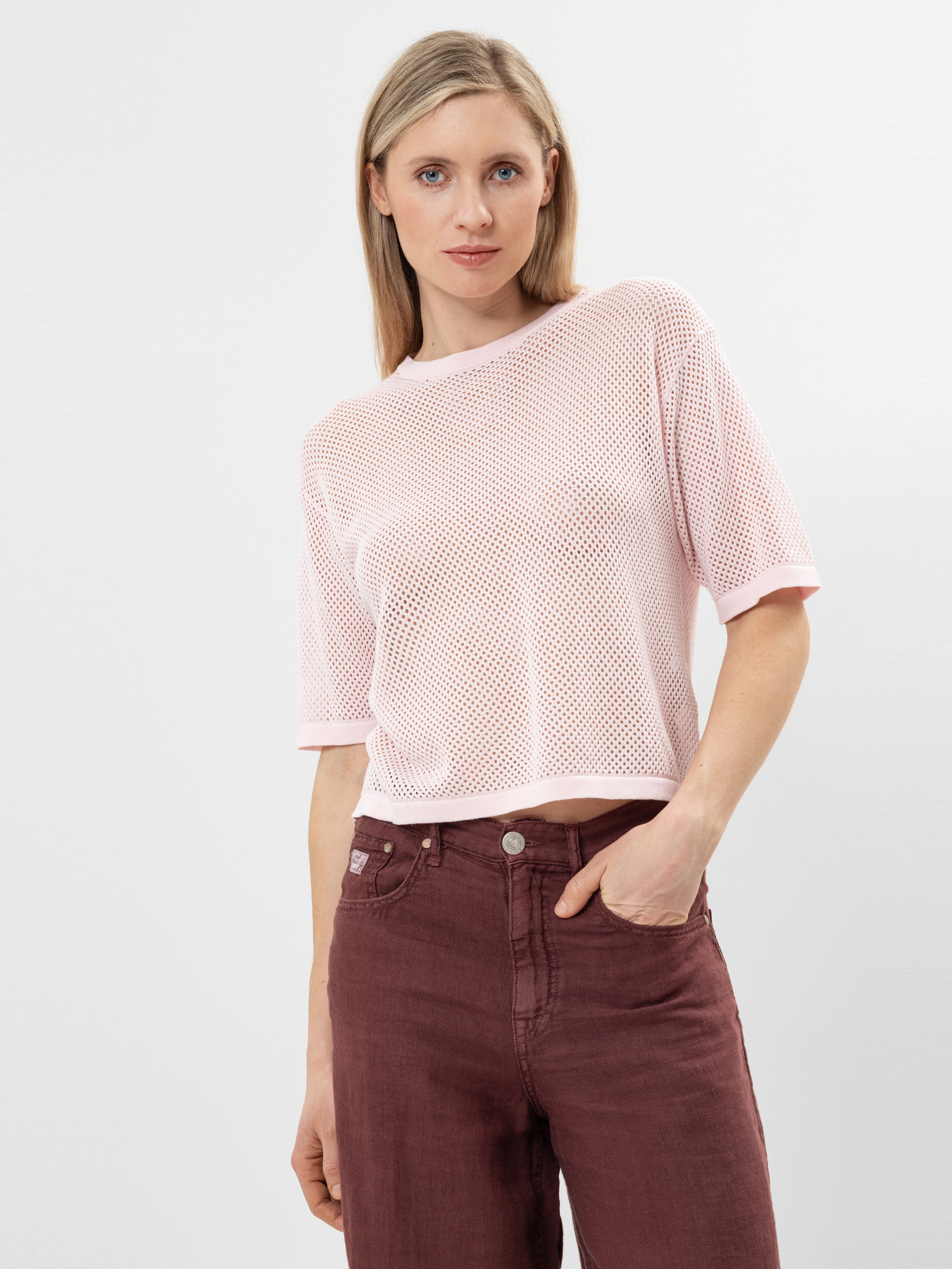 Eine Frau mit glattem blondem Haar, die ein blassrosa Netzoberteil mit kurzen Ärmeln und eine hoch sitzende kastanienbraune Jeans trägt, steht vor einem schlichten weißen Hintergrund und hat eine Hand in der Tasche.