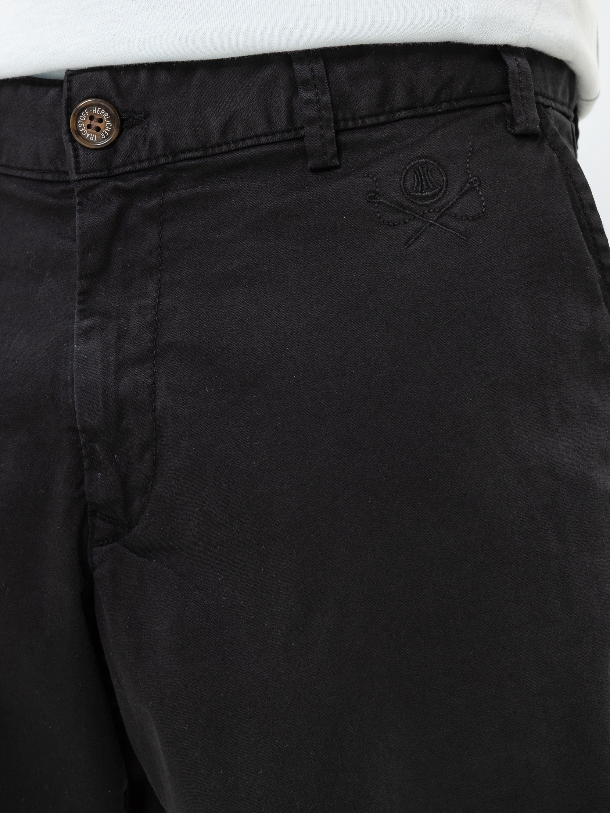 Nahaufnahme einer schwarzen Hose mit einem braunen Knopf, schwarzen Nähten und einem gestickten Logo mit einem Basketball und zwei gekreuzten Schlägern in der Nähe der Vordertasche. An der Taille ist der Rand eines weißen Hemdes zu sehen.
