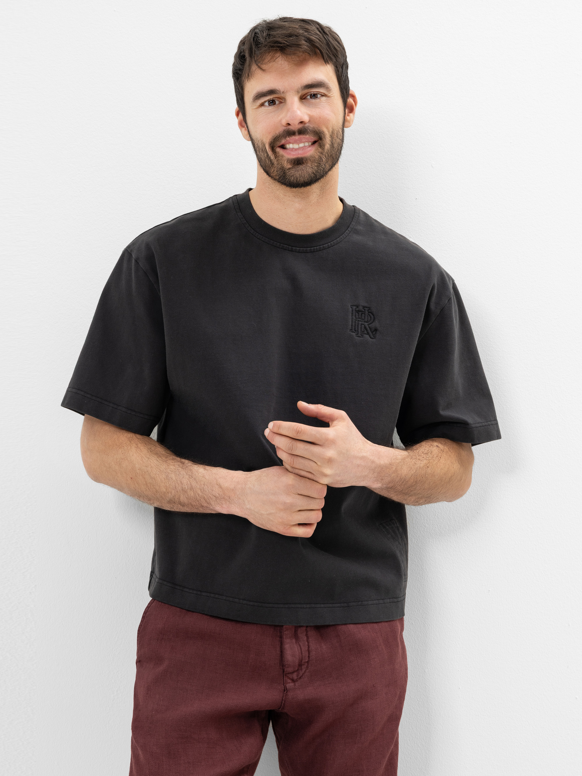 Ein Mann mit kurzen dunklen Haaren und einem Bart steht lächelnd vor einer weißen Wand. Er trägt ein lockeres schwarzes T-Shirt und eine kastanienbraune Hose, die eine Hand hält die andere vor sich.