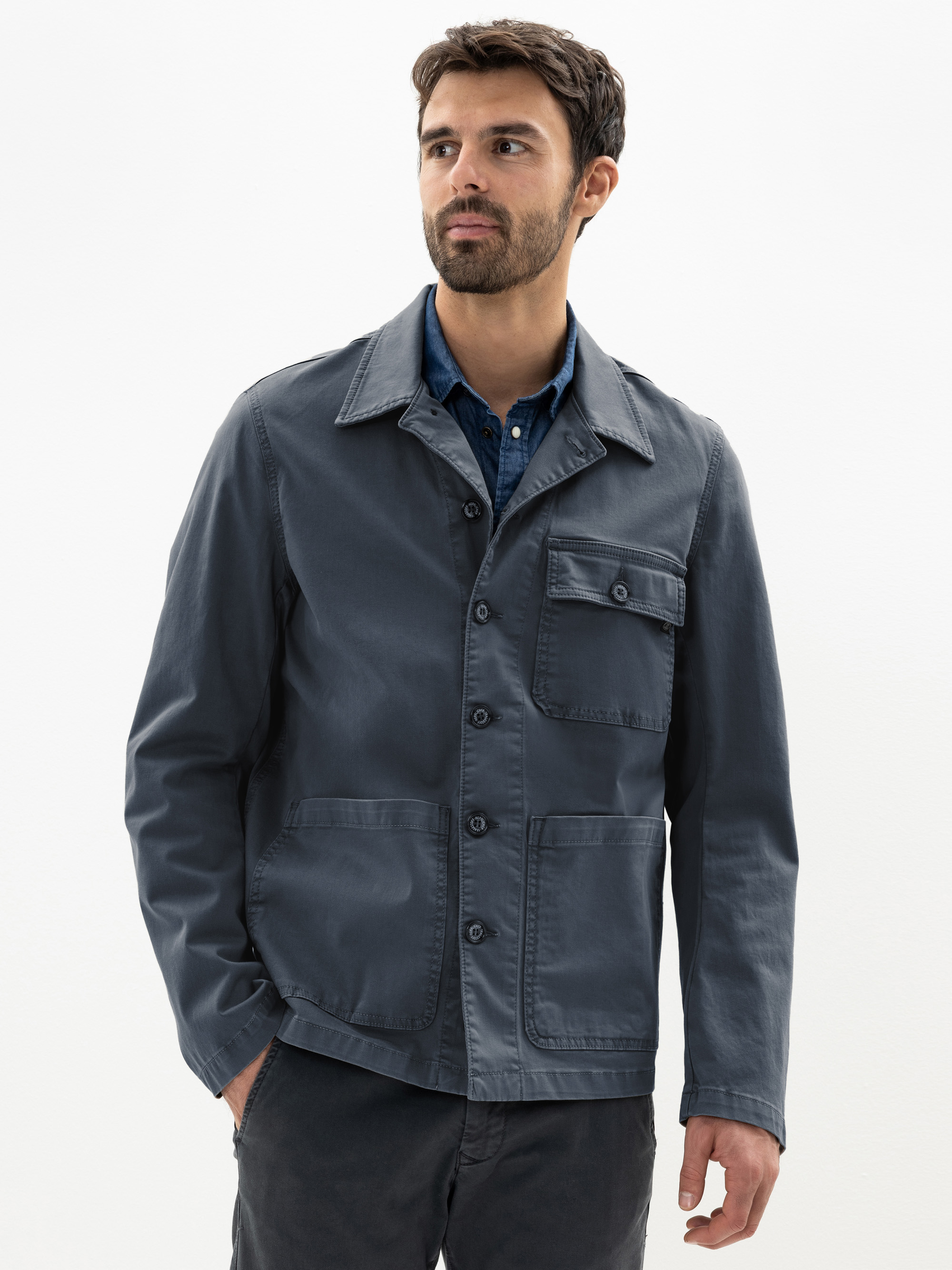 Ein Mann mit kurzen dunklen Haaren und einem Bart trägt eine blaue Jacke mit Knöpfen über einem blauen Hemd. Er steht mit einer Hand in der Tasche vor einem schlichten weißen Hintergrund und schaut leicht zur Seite.