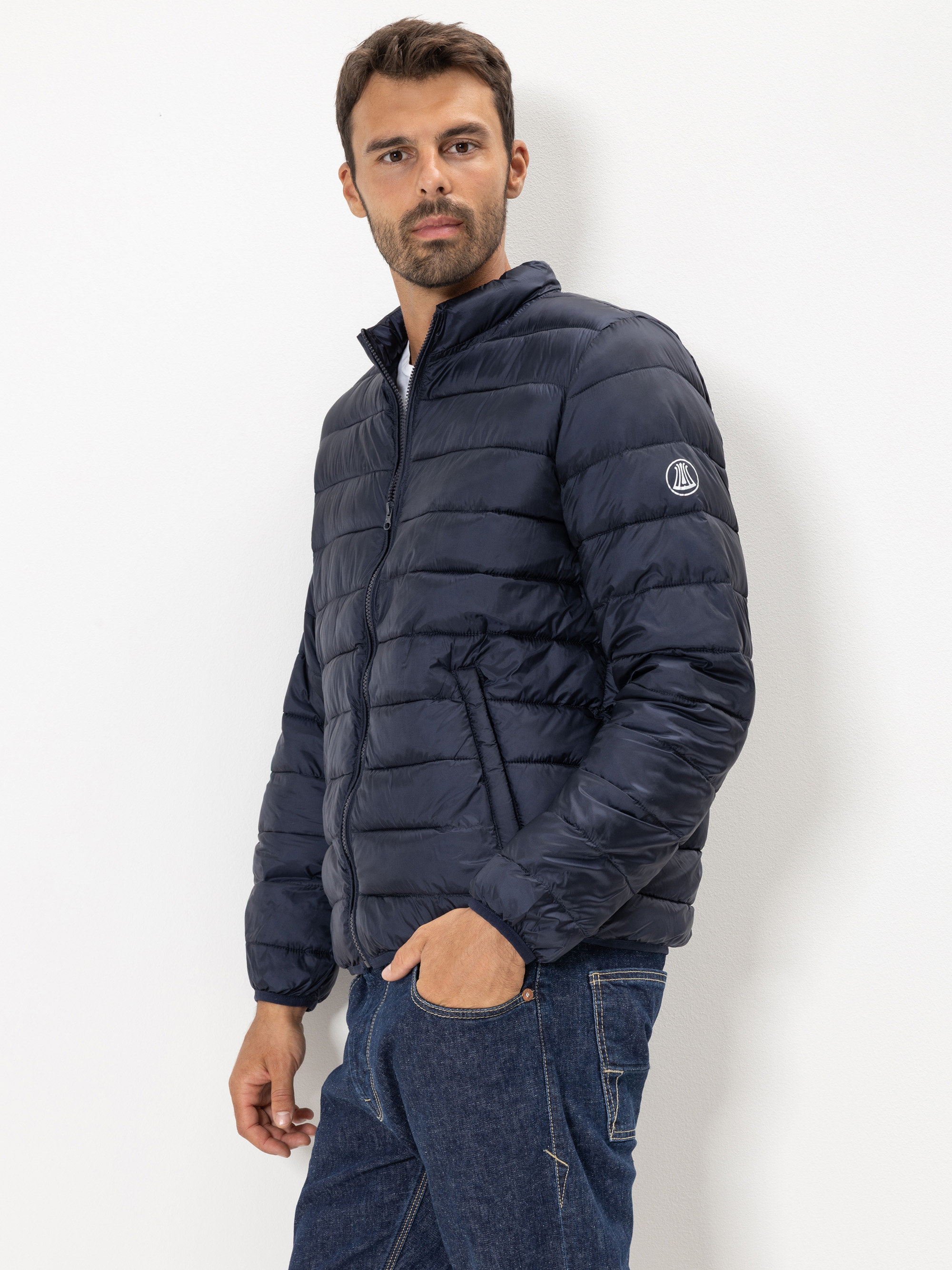 Ein Mann mit kurzen dunklen Haaren und einem Bart trägt eine marineblaue Steppjacke und blaue Jeans und steht mit einer Hand in der Tasche vor einem schlichten weißen Hintergrund.