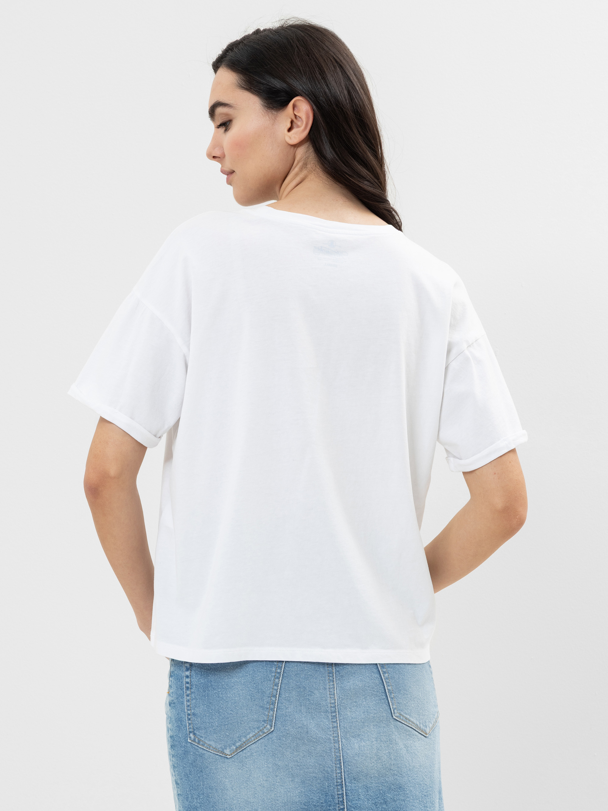 Eine Frau mit langen dunklen Haaren steht mit dem Rücken zur Kamera, trägt ein lockeres weißes T-Shirt und einen hellblauen Jeansrock vor einem schlichten hellen Hintergrund.
