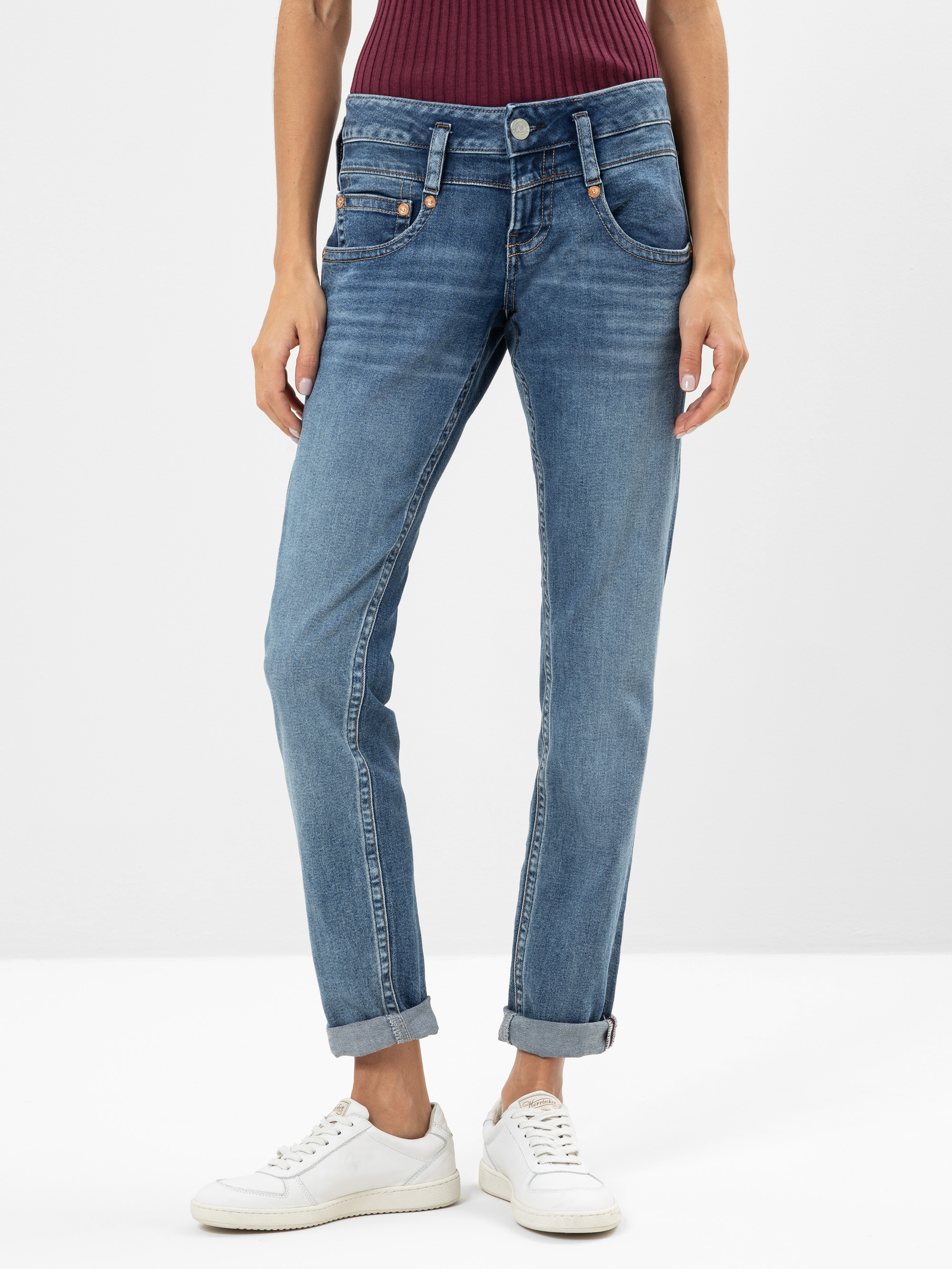 Person in blauen Jeans mit gekrempelten Manschetten, einem gerippten weinroten Oberteil und weißen Turnschuhen, die vor einem schlichten hellen Hintergrund steht. Nur der untere Teil des Oberkörpers und die Beine sind sichtbar.