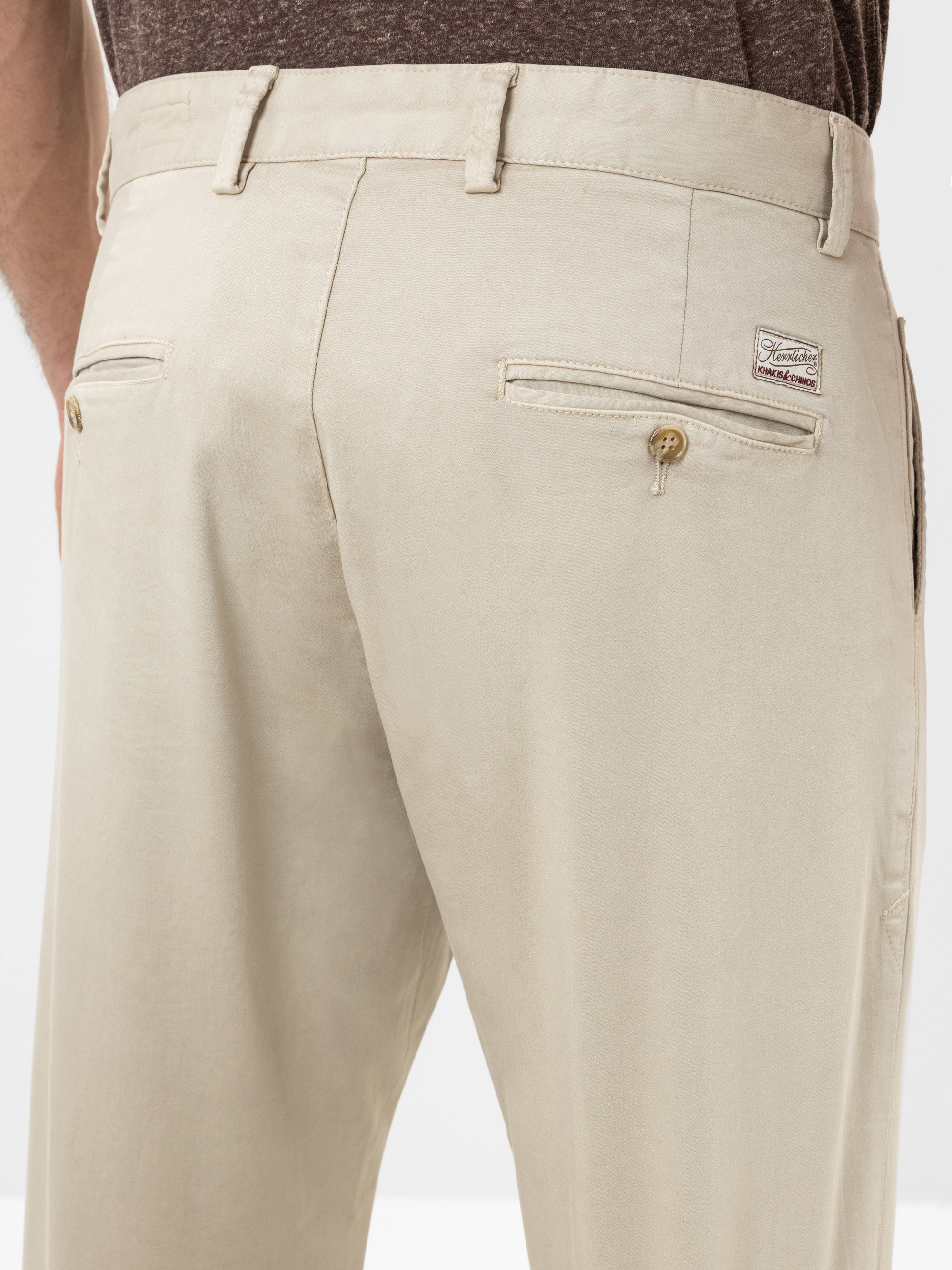 Nahaufnahme des Rückens einer Person, die eine beigefarbene Anzughose mit geknöpften Gesäßtaschen und einem kleinen Markenetikett aus Stoff trägt, dazu ein braunes Hemd, das in die Hose gesteckt wird.