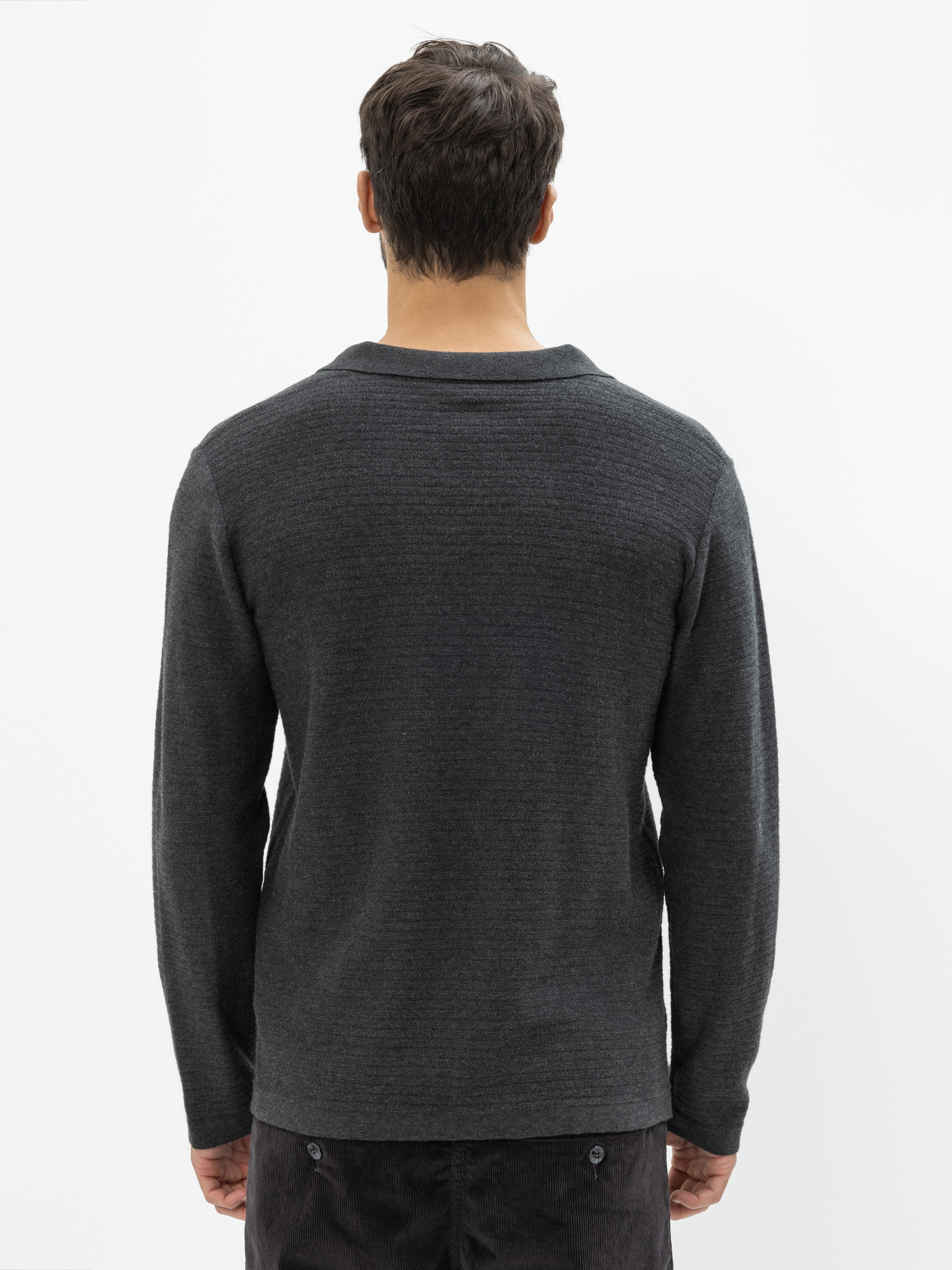 Ein Mann steht mit dem Rücken zur Kamera, trägt einen dunkelgrauen langärmeligen Pullover und eine dunkle Hose vor einem schlichten weißen Hintergrund.