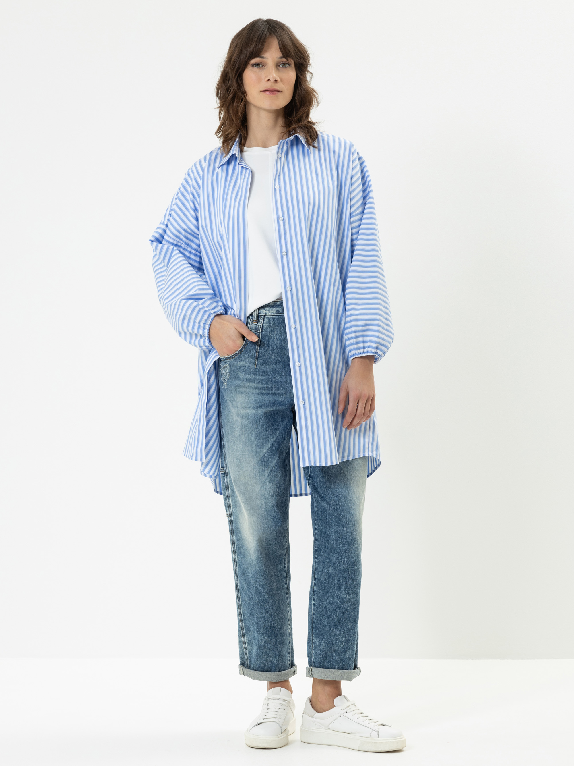 Eine Frau steht vor einem schlichten weißen Hintergrund und trägt ein lockeres, blau-weiß gestreiftes Oversized-Shirt, ein weißes T-Shirt, blaue Jeans und weiße Turnschuhe. Sie hat schulterlanges gewelltes Haar und eine entspannte Pose.