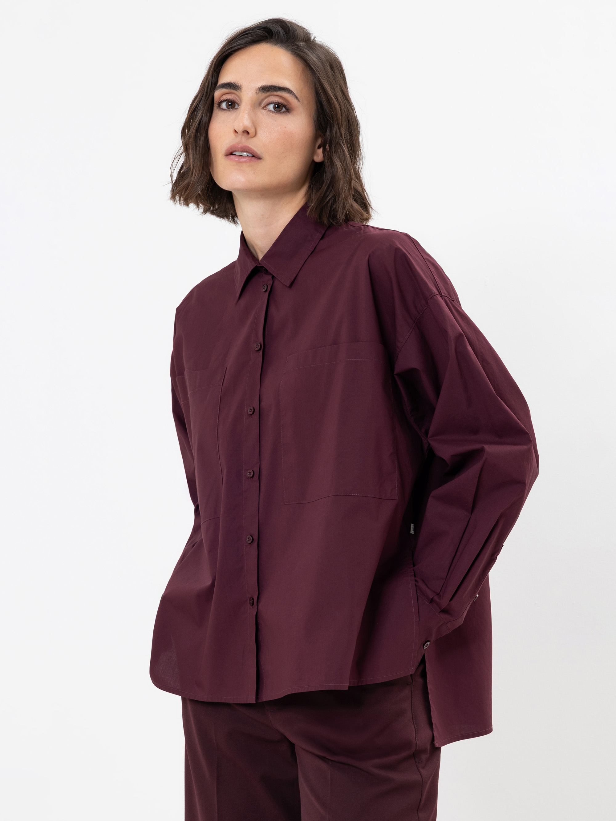 Eine Frau mit kurzen braunen Haaren, die ein locker sitzendes, langärmeliges kastanienbraunes Button-up-Hemd und eine passende kastanienbraune Hose trägt, steht vor einem schlichten weißen Hintergrund.