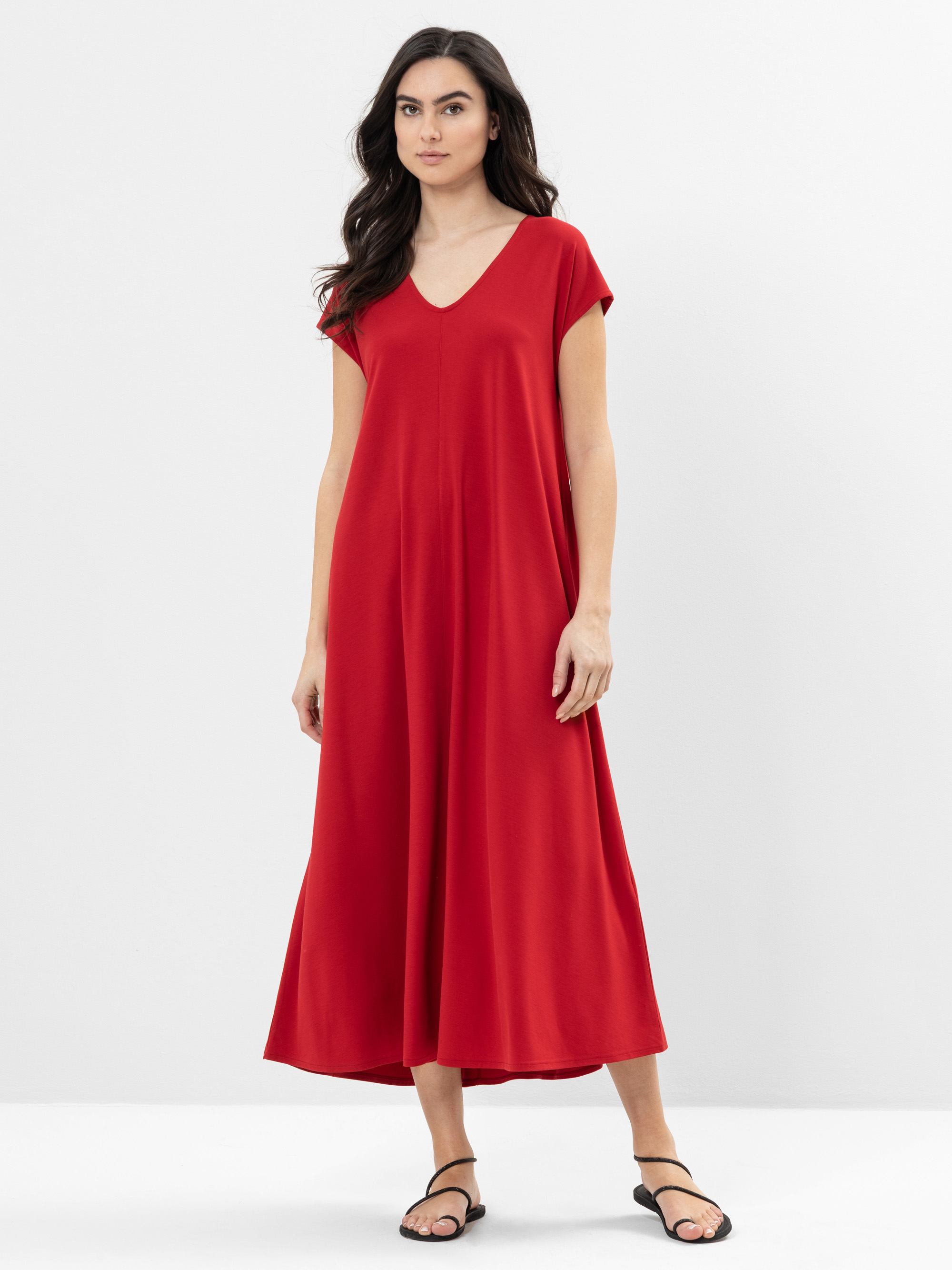Eine Frau mit langen dunklen Haaren trägt ein locker sitzendes, kurzärmeliges, knöchellanges rotes Kleid mit V-Ausschnitt und schwarze Sandalen und steht vor einem schlichten weißen Hintergrund.