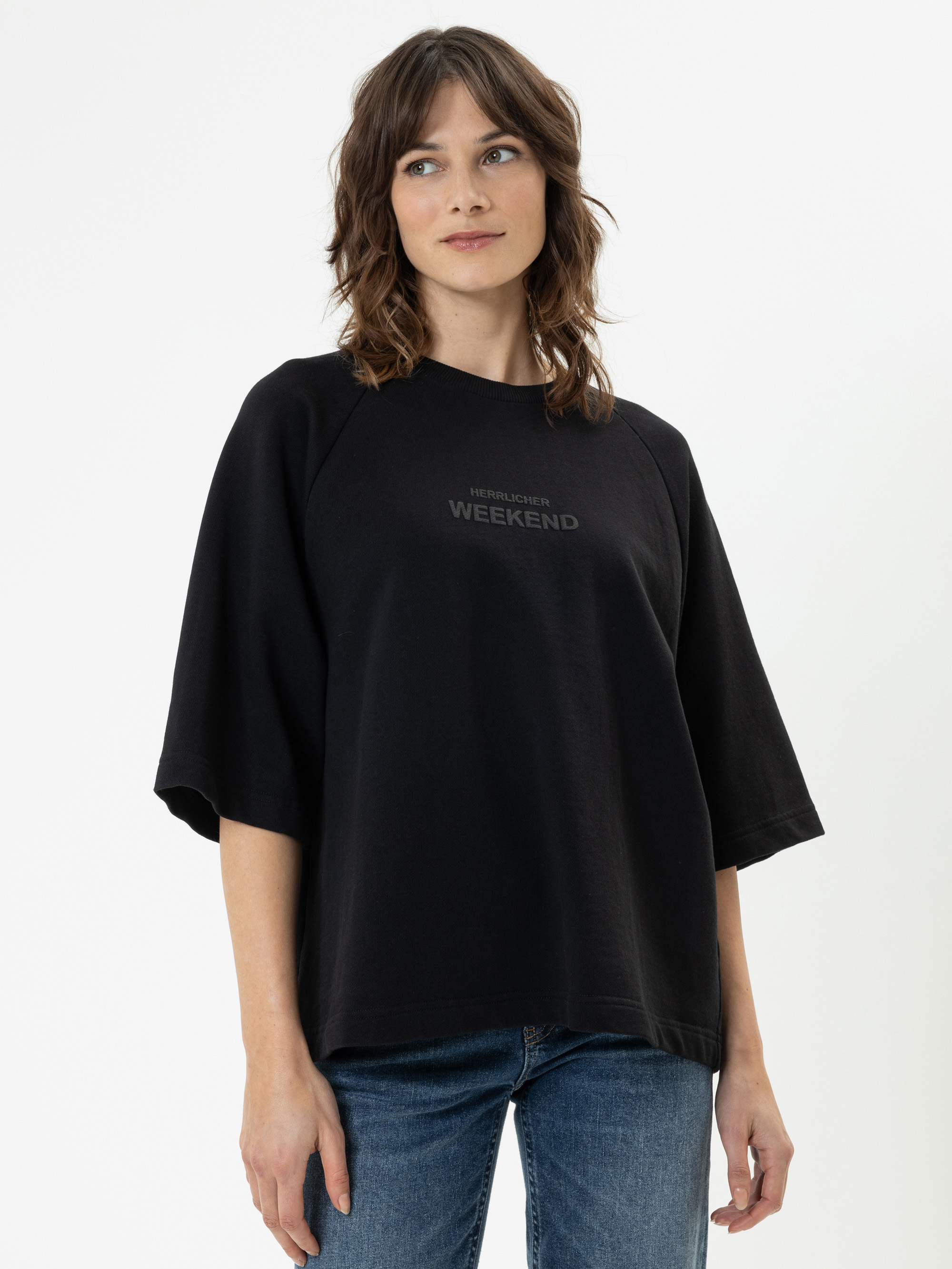Eine Frau mit gewelltem braunem Haar, die ein locker sitzendes schwarzes T-Shirt mit der Aufschrift WEEKEND auf der Vorderseite und blaue Jeans trägt, steht vor einem schlichten weißen Hintergrund.