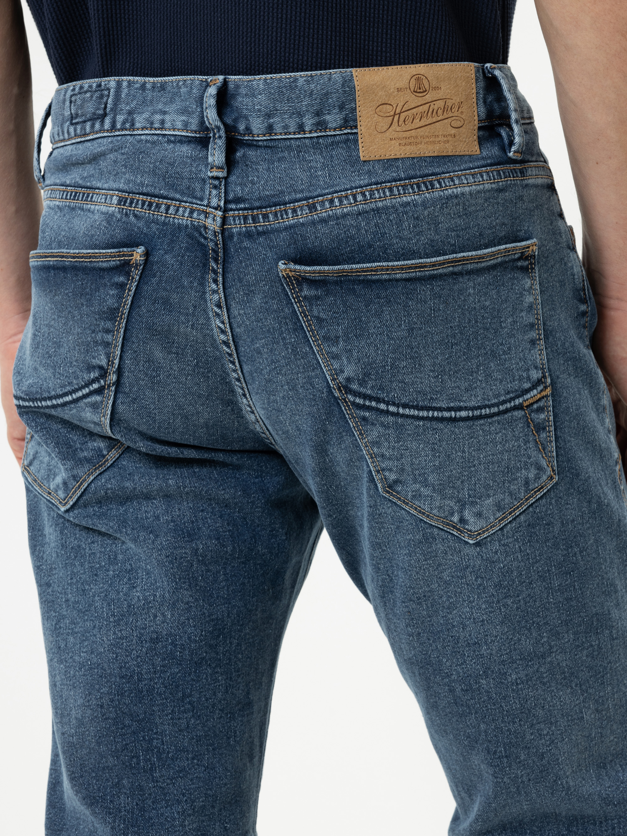 Nahaufnahme des Rückens einer Person, die eine blaue Denim-Jeans mit großen Gesäßtaschen und einem braunen Lederaufnäher mit dem Markennamen Heritage am Bund trägt.