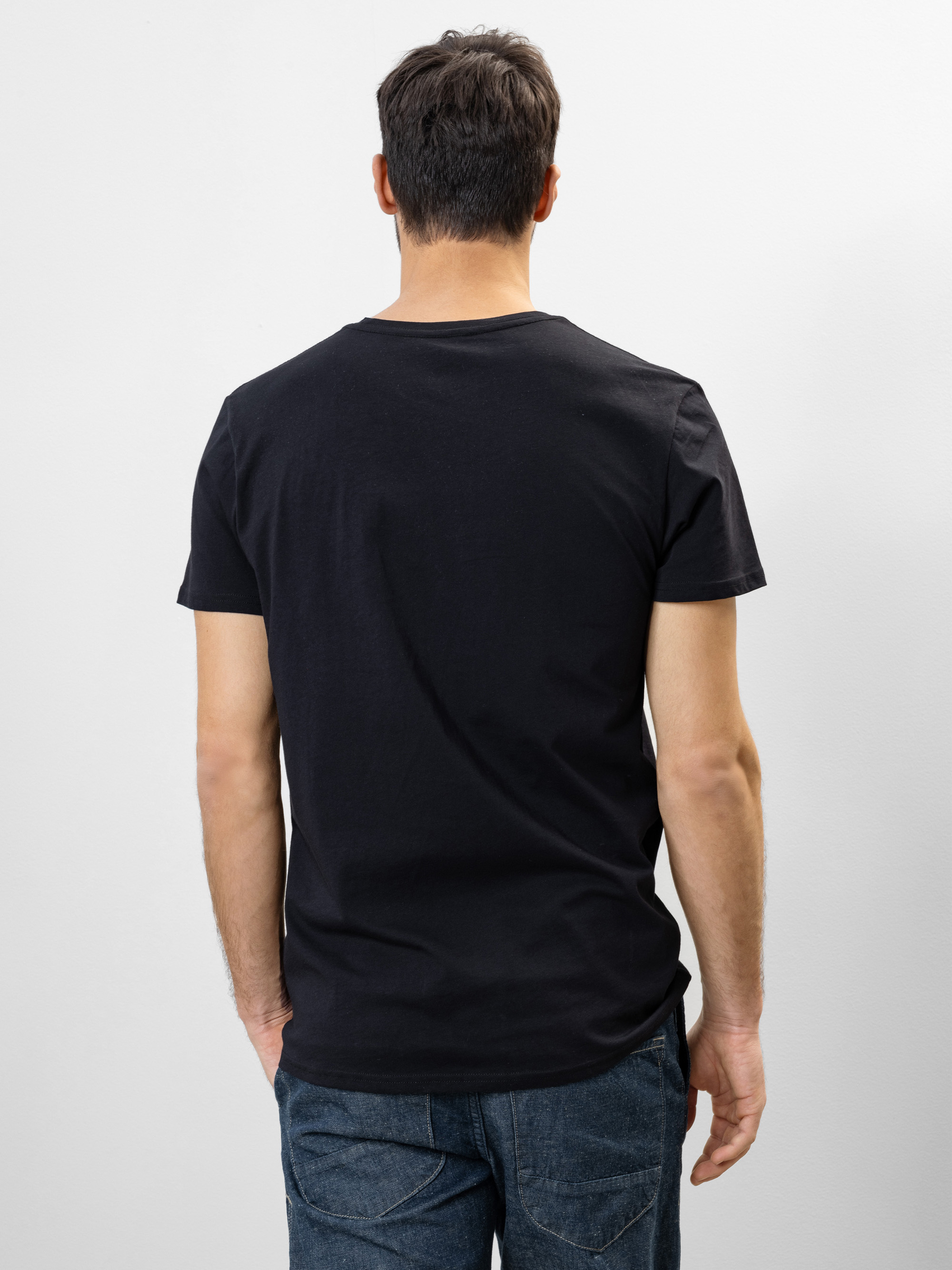 Ein Mann mit kurzen braunen Haaren steht mit dem Rücken zur Kamera, trägt ein schlichtes schwarzes T-Shirt und blaue Jeans vor einem schlichten weißen Hintergrund.