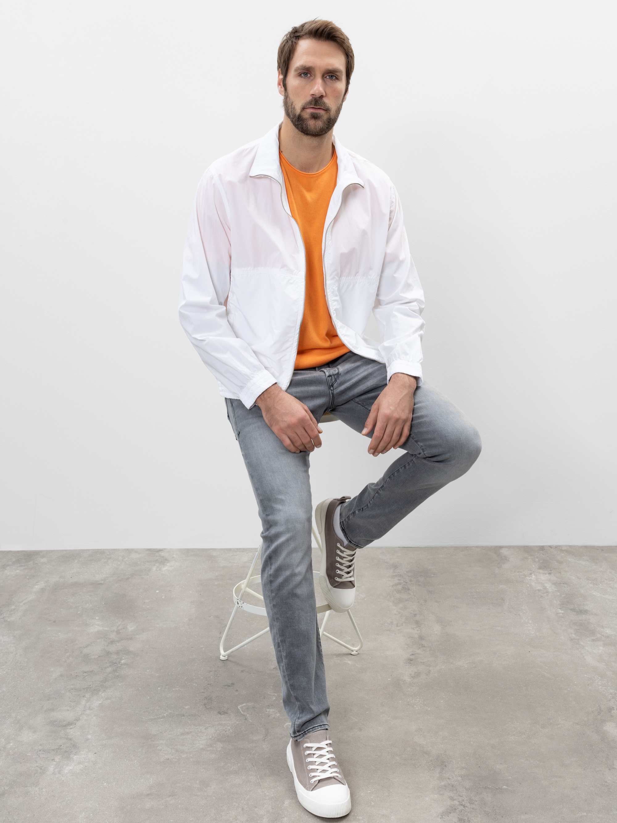 Ein Mann mit kurzen braunen Haaren und einem Bart sitzt auf einem weißen Hocker. Er trägt eine weiße Jacke, ein orangefarbenes Hemd, hellgraue Jeans und graue Turnschuhe. Der Hintergrund ist schlicht und minimalistisch mit einem Betonboden und einer we