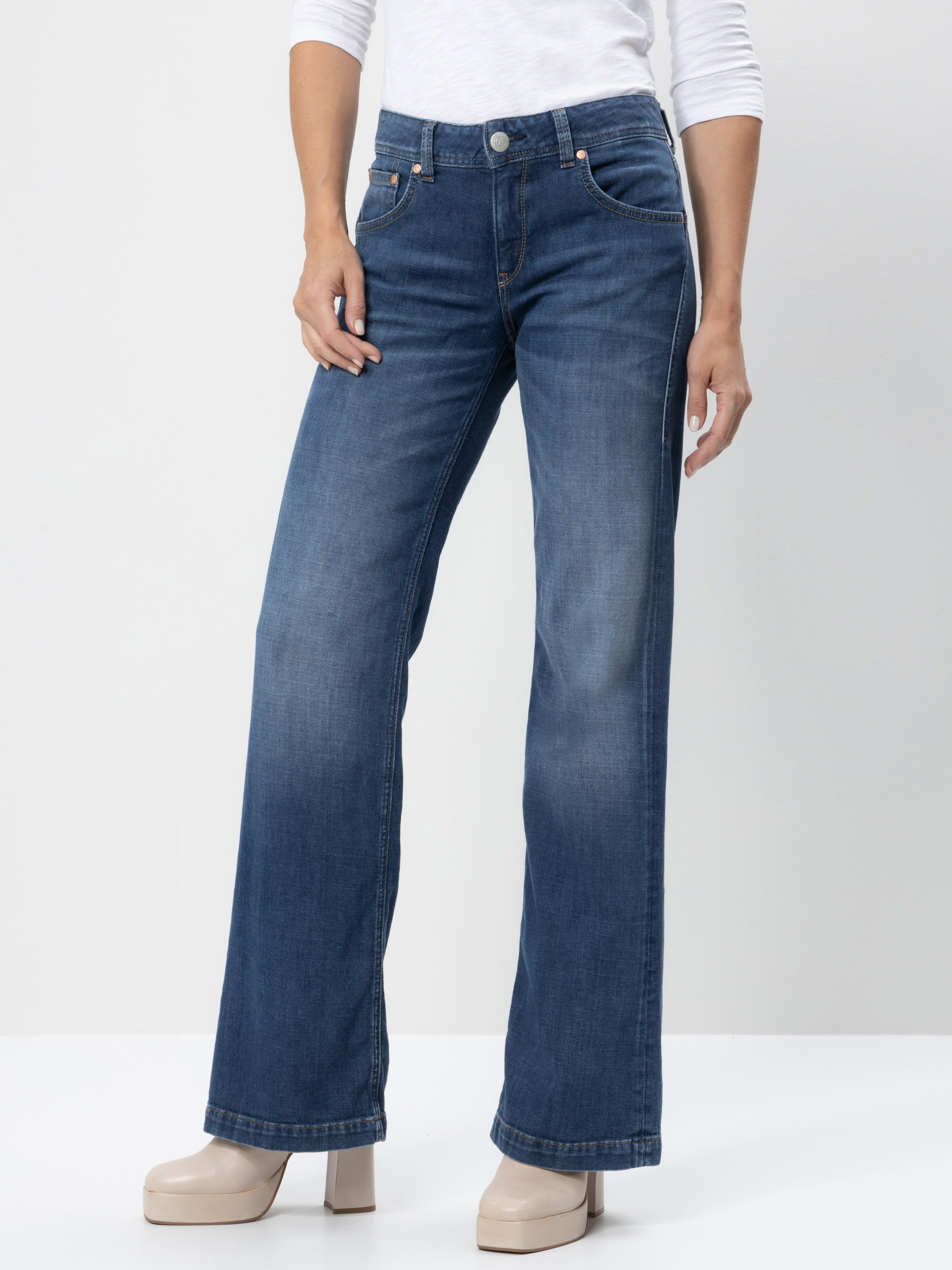 Eine Person, die eine blaue Jeans mit weitem Bein, ein weißes langärmeliges Oberteil und beigefarbene Plateauschuhe trägt, steht vor einem schlichten hellen Hintergrund. Das Gesicht der Person ist auf dem Bild nicht zu sehen.