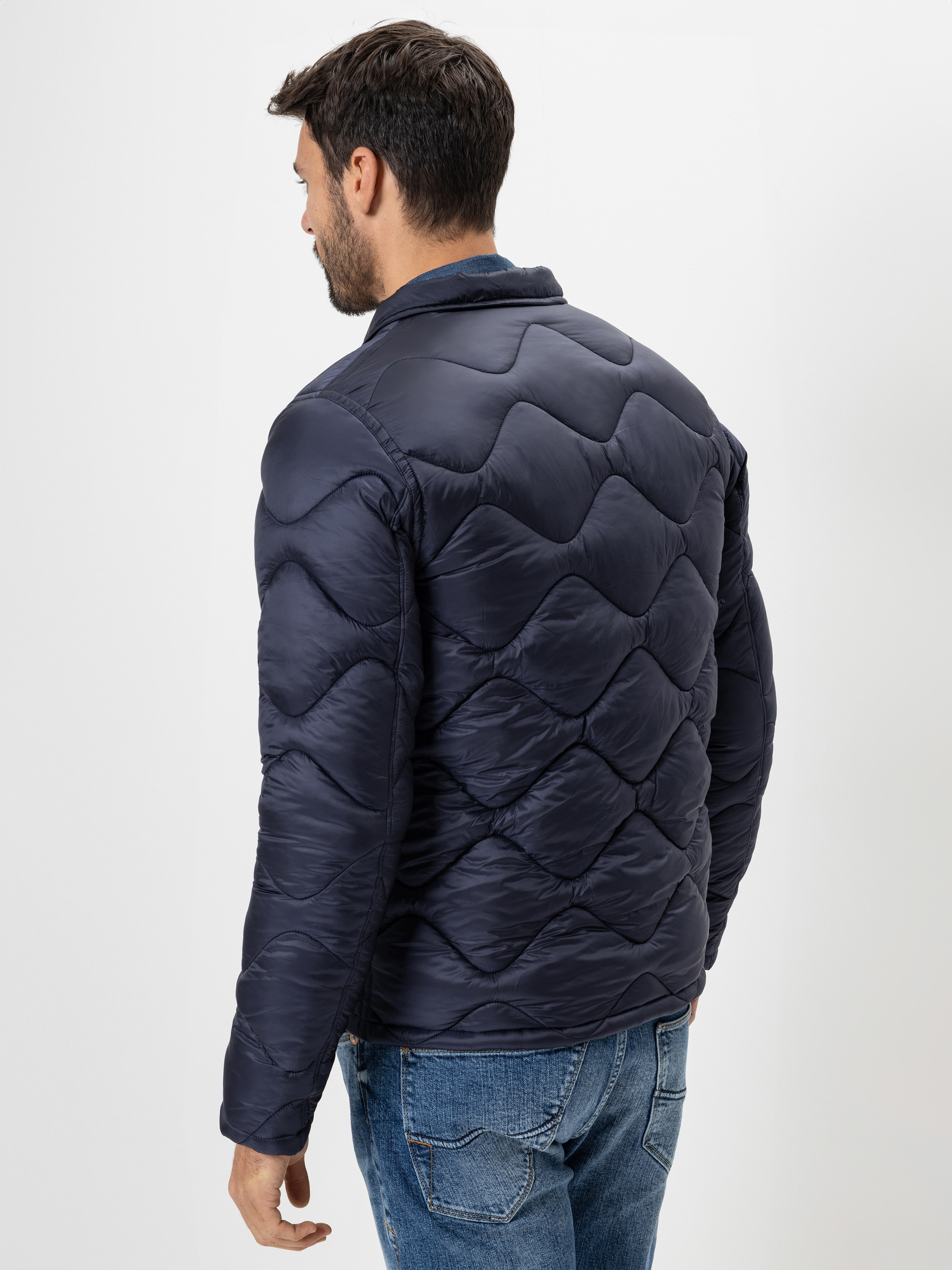 Ein Mann mit kurzen dunklen Haaren, von hinten gesehen, trägt eine marineblaue Steppjacke und blaue Jeans vor einem schlichten weißen Hintergrund.