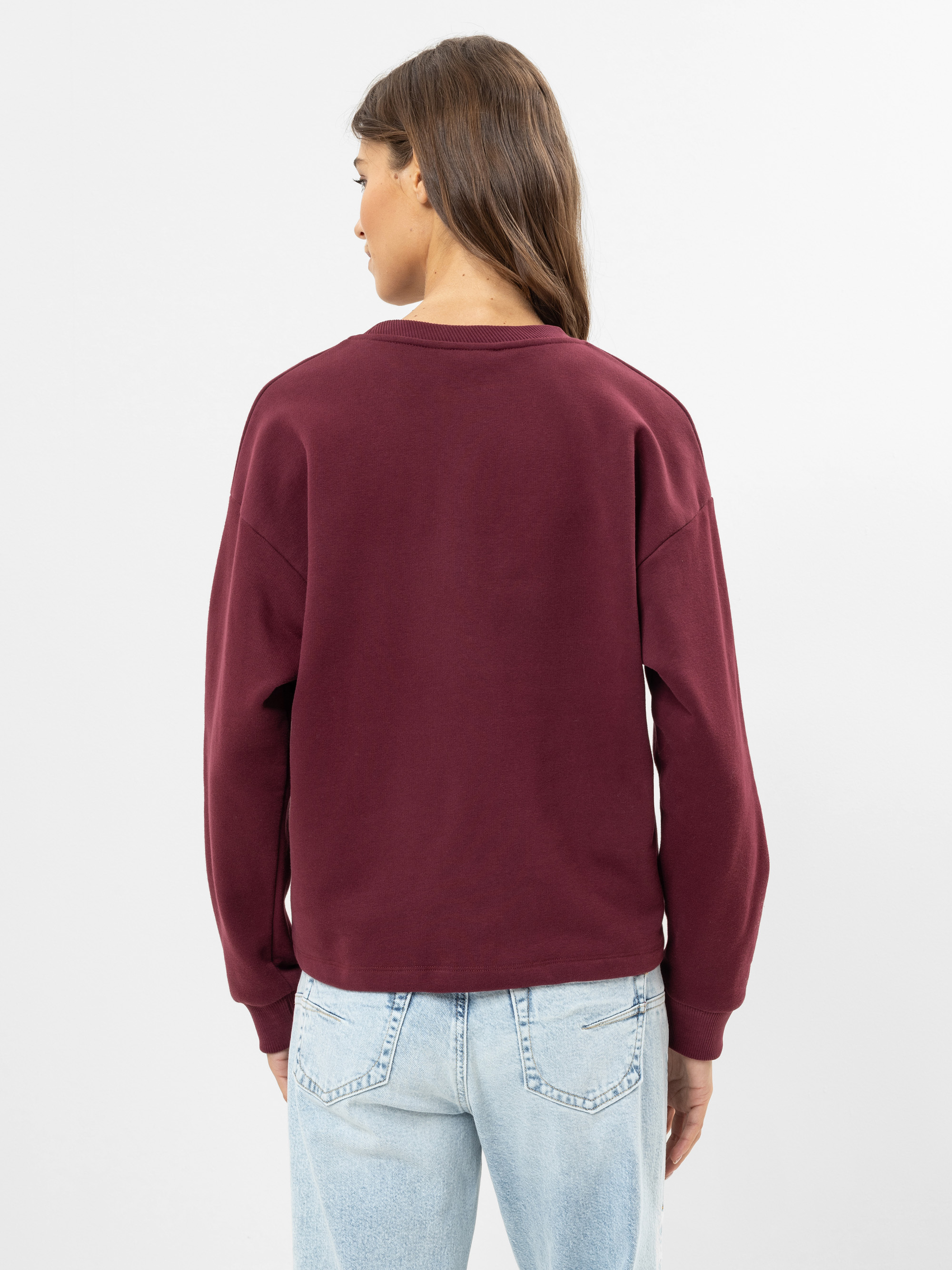 Eine Person mit langen braunen Haaren, die ein kastanienbraunes Sweatshirt und hellblaue Jeans trägt, steht mit dem Gesicht zur Kamera vor einem schlichten weißen Hintergrund.