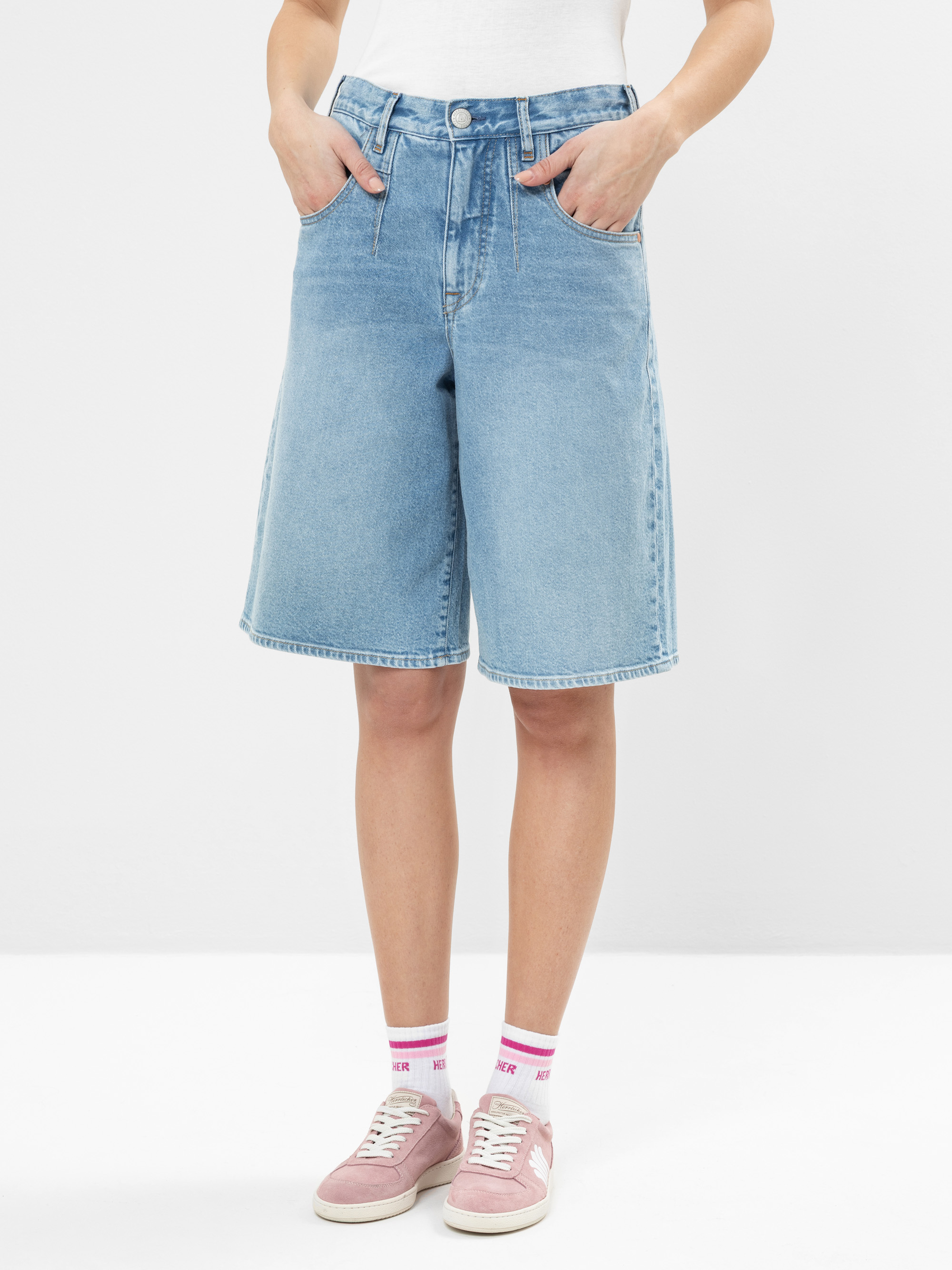 Eine Person in hellblauen knielangen Jeans-Shorts, einem weißen Oberteil, rosa-weißen Socken und rosa Turnschuhen steht mit den Händen in den Taschen vor einem schlichten weißen Hintergrund.