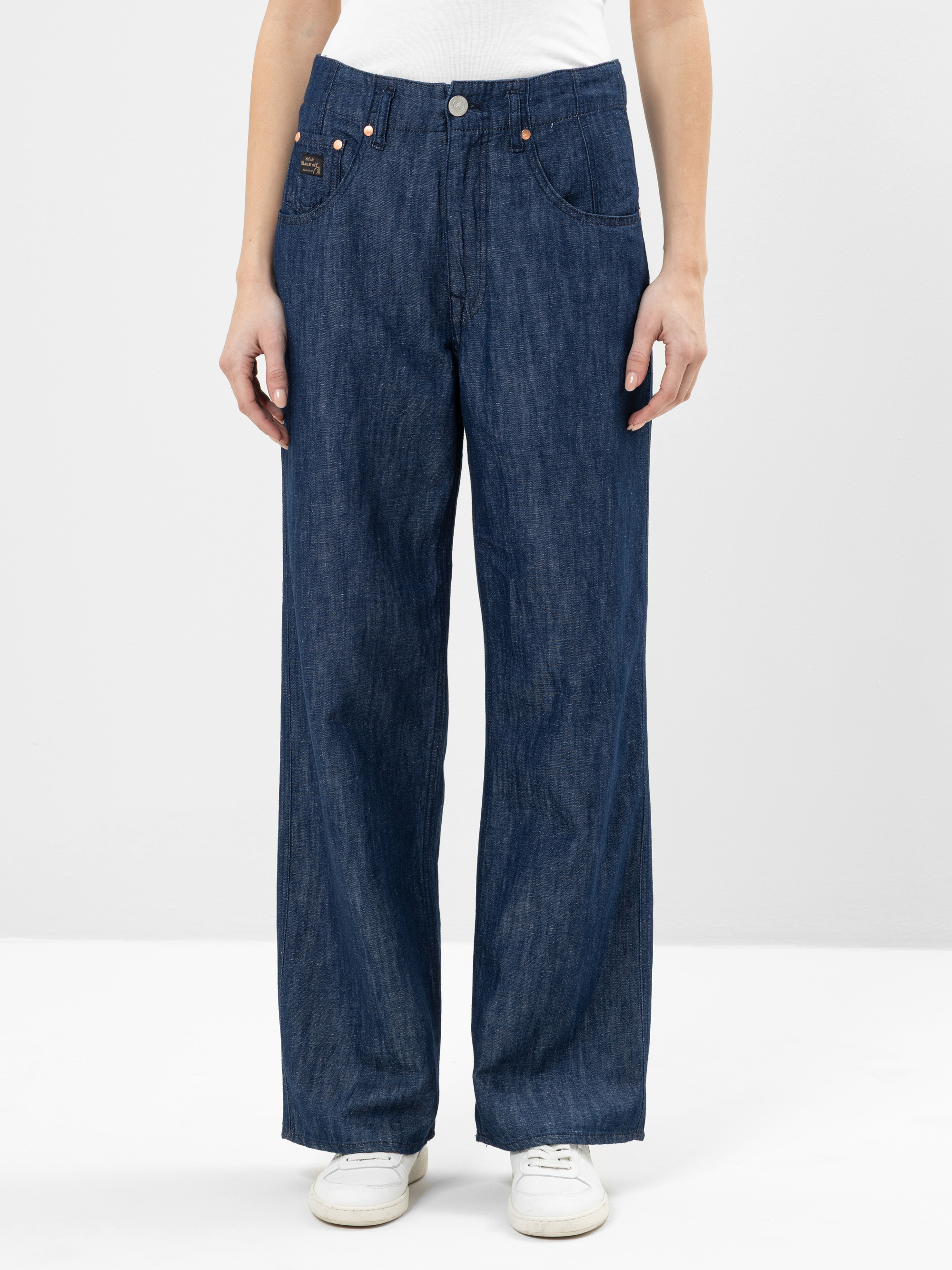 Eine Person, die eine hochgekrempelte, weit geschnittene Bluejeans und weiße Turnschuhe trägt, steht vor einem schlichten weißen Hintergrund und hält die Hände locker an der Seite.