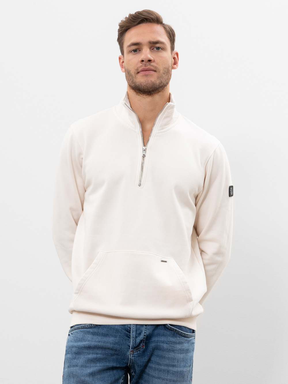 Ein Mann mit kurzen braunen Haaren und gestutztem Bart trägt einen hellen Pullover mit viertelhohem Reißverschluss und blaue Jeans und steht mit den Händen auf dem Rücken vor einem schlichten weißen Hintergrund.
