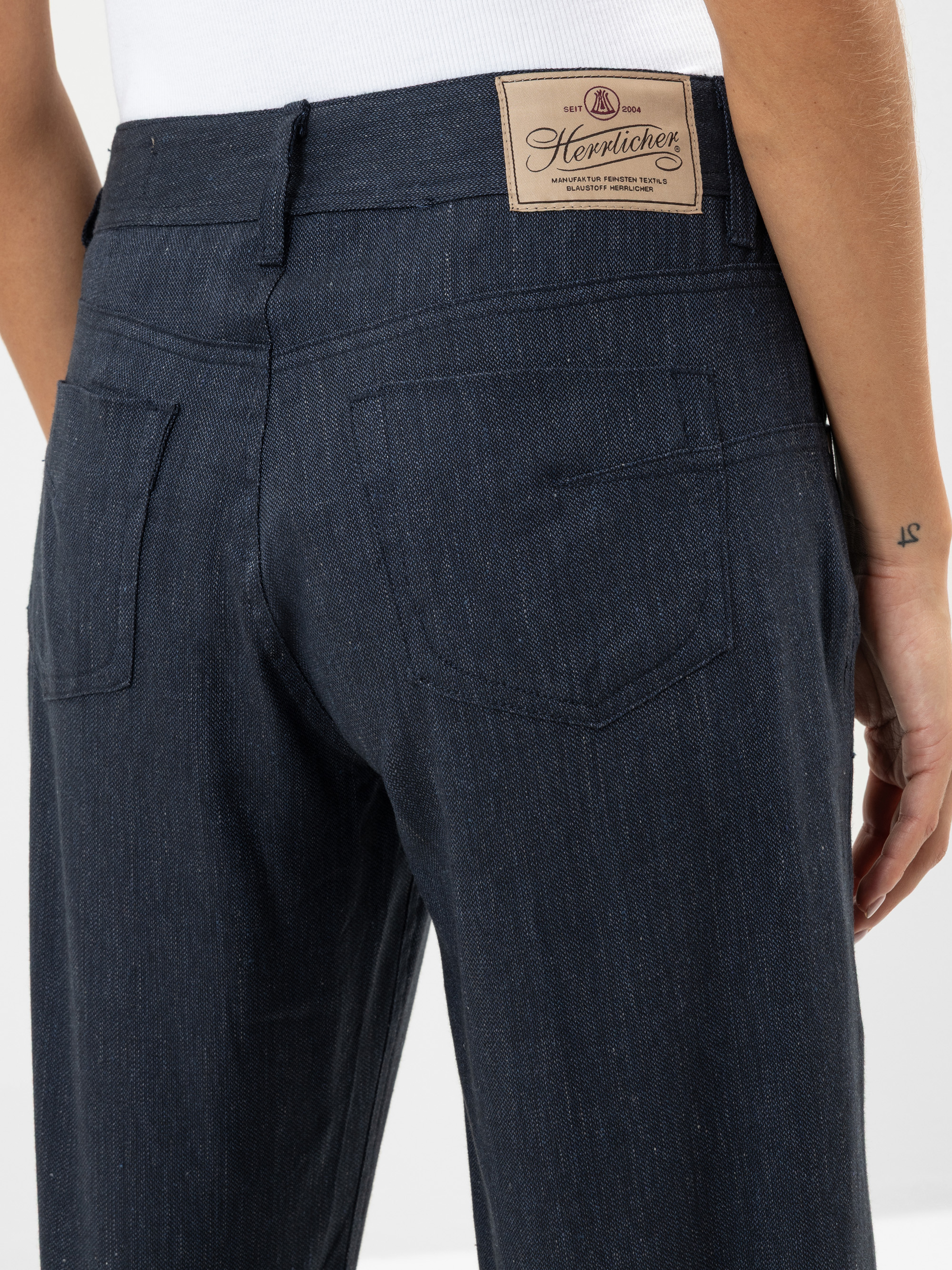 Nahaufnahme einer Person, die eine dunkelblaue Jeans trägt, die Gesäßtaschen und ein beigefarbenes Etikett der Marke Herzlichen am Bund zeigt. Die Person hat eine kleine 12-Tätowierung am Handgelenk.