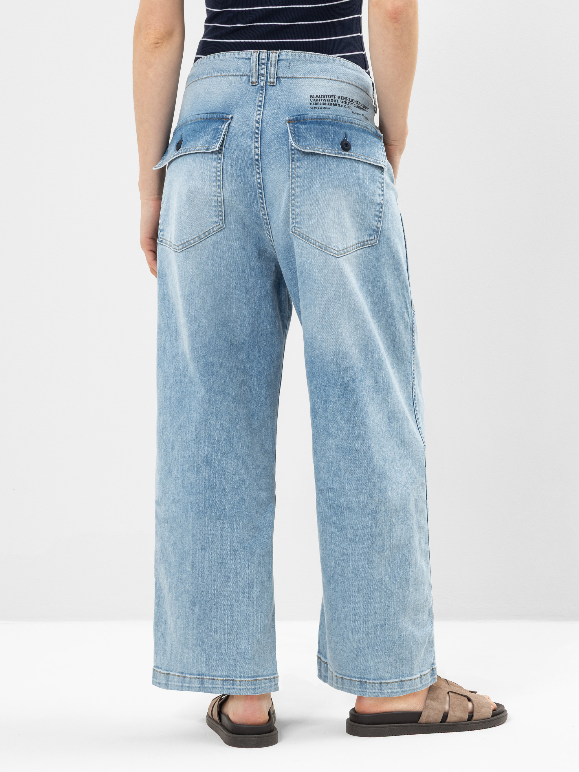 Person in hellblauen, weit geschnittenen Jeans mit großen Gesäßtaschen und einem gestreiften marineblauen Oberteil, stehend auf weißem Hintergrund. Nur die untere Hälfte des Körpers ist sichtbar; die Person trägt außerdem braune Sandalen.