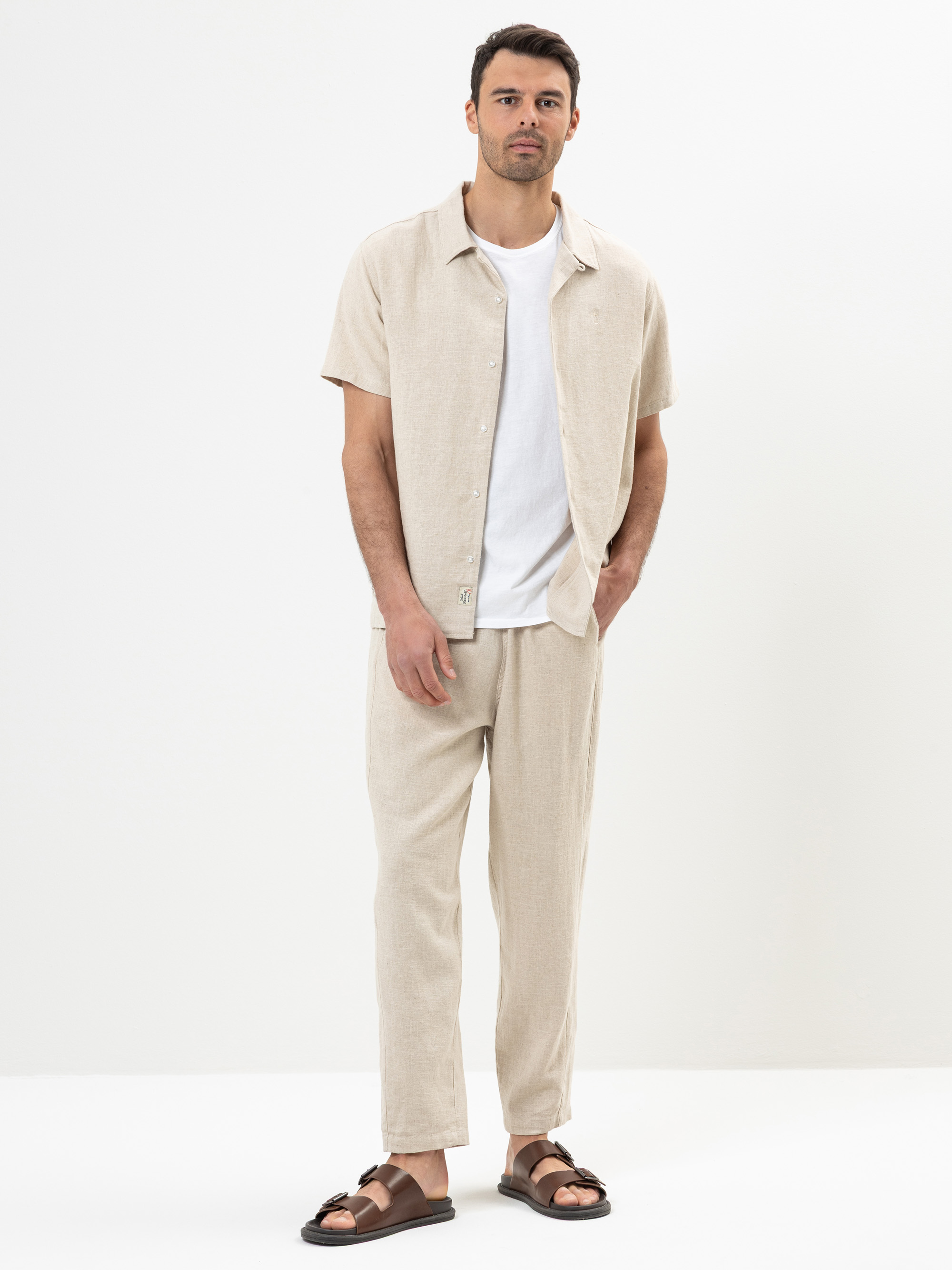 Ein Mann steht vor einem schlichten weißen Hintergrund und trägt ein beigefarbenes kurzärmeliges Hemd über einem weißen T-Shirt, eine passende beigefarbene Hose und braune Sandalen. Er hat eine Hand in der Tasche und einen neutralen Gesichtsausdruck.