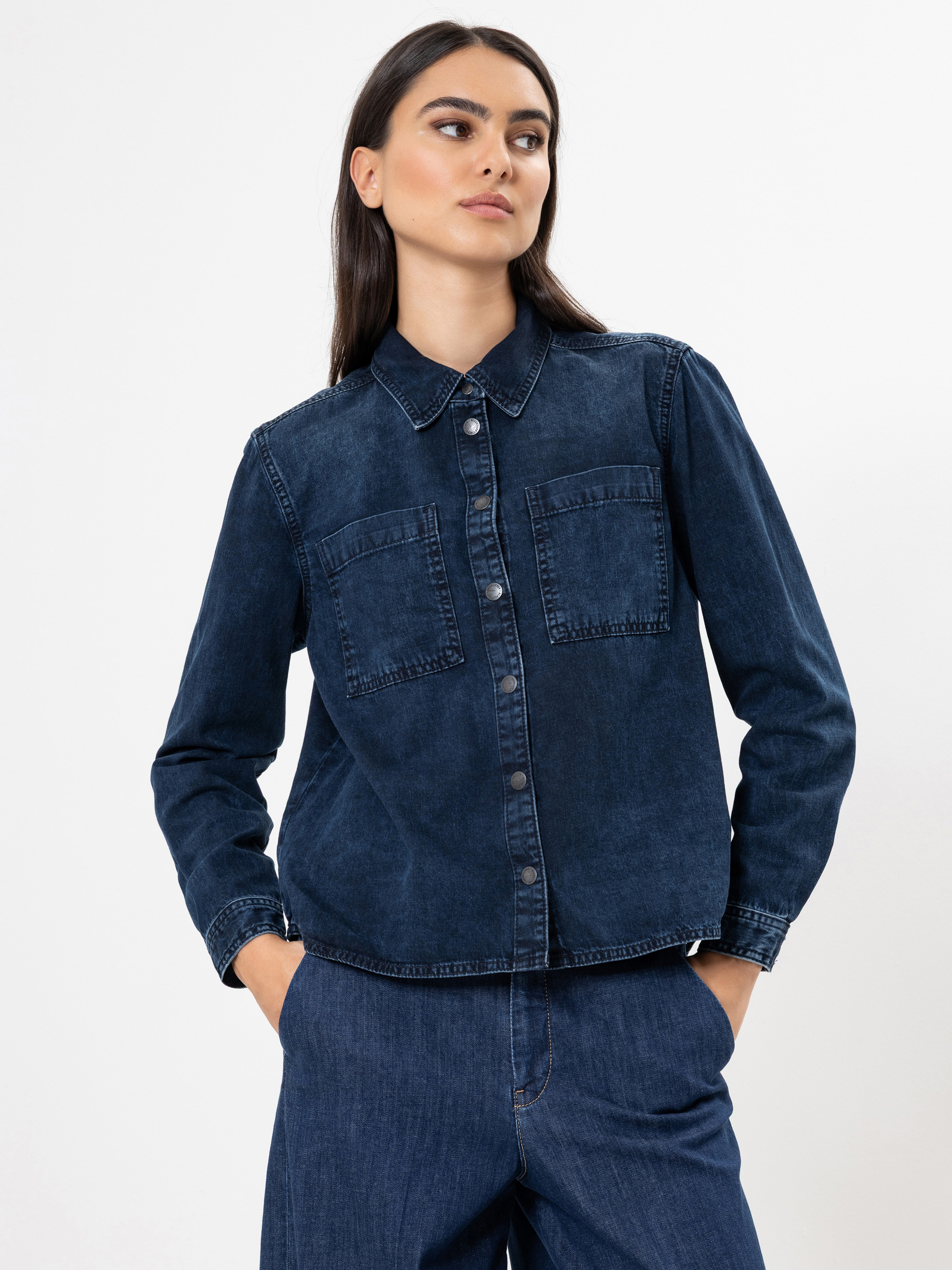 Eine Frau mit langen dunklen Haaren trägt ein dunkelblaues Jeanshemd zum Aufknöpfen mit Vordertaschen und eine passende Jeanshose. Sie steht mit den Händen in den Taschen vor einem schlichten weißen Hintergrund.