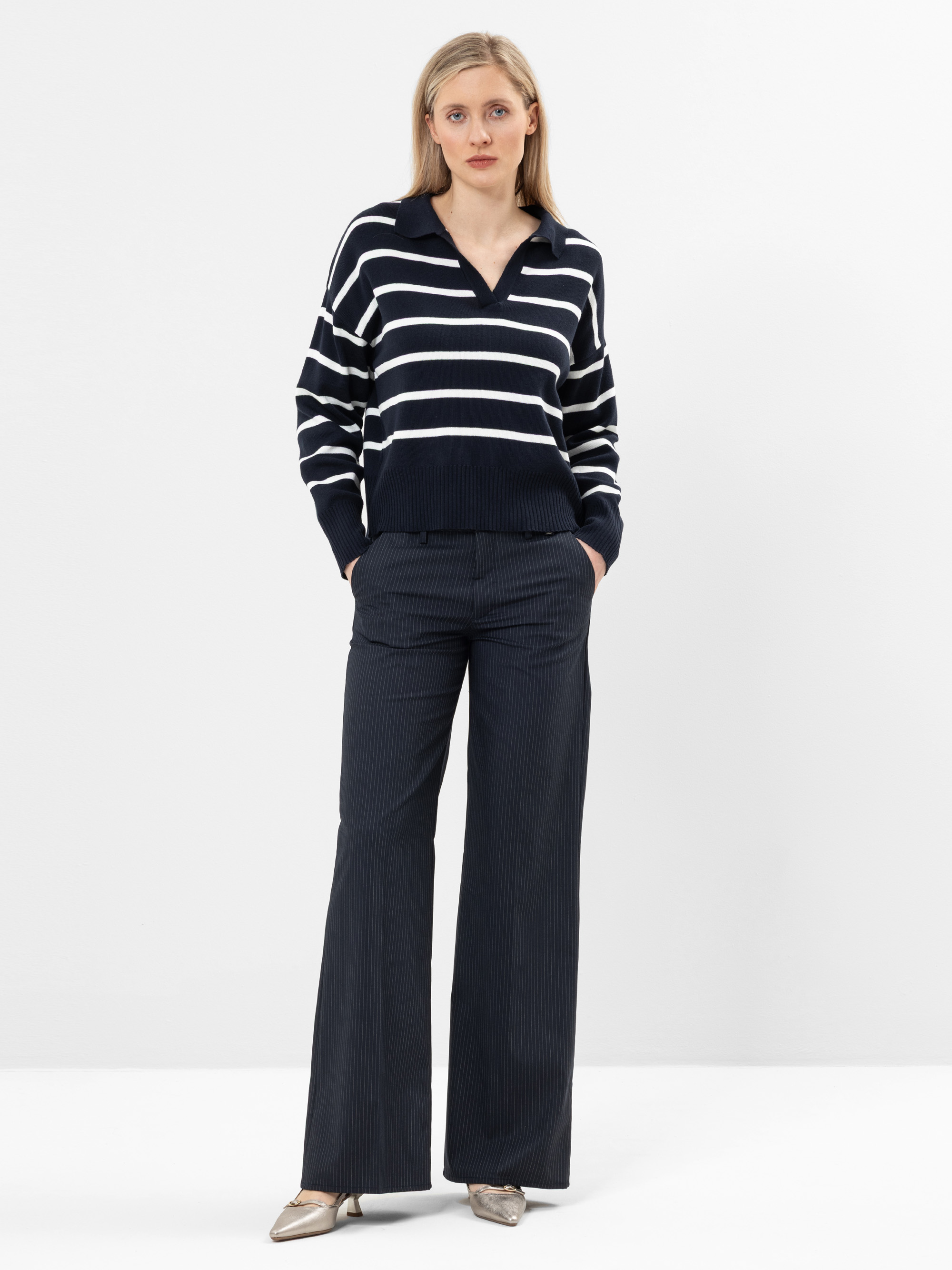 Eine Frau mit blondem Haar steht vor einem weißen Hintergrund und trägt einen marineblauen Pullover mit weißen Querstreifen, eine marineblaue Hose mit weitem Bein und Metallic-Schuhe. Sie hat die Hände in den Taschen und blickt in die Kamera.