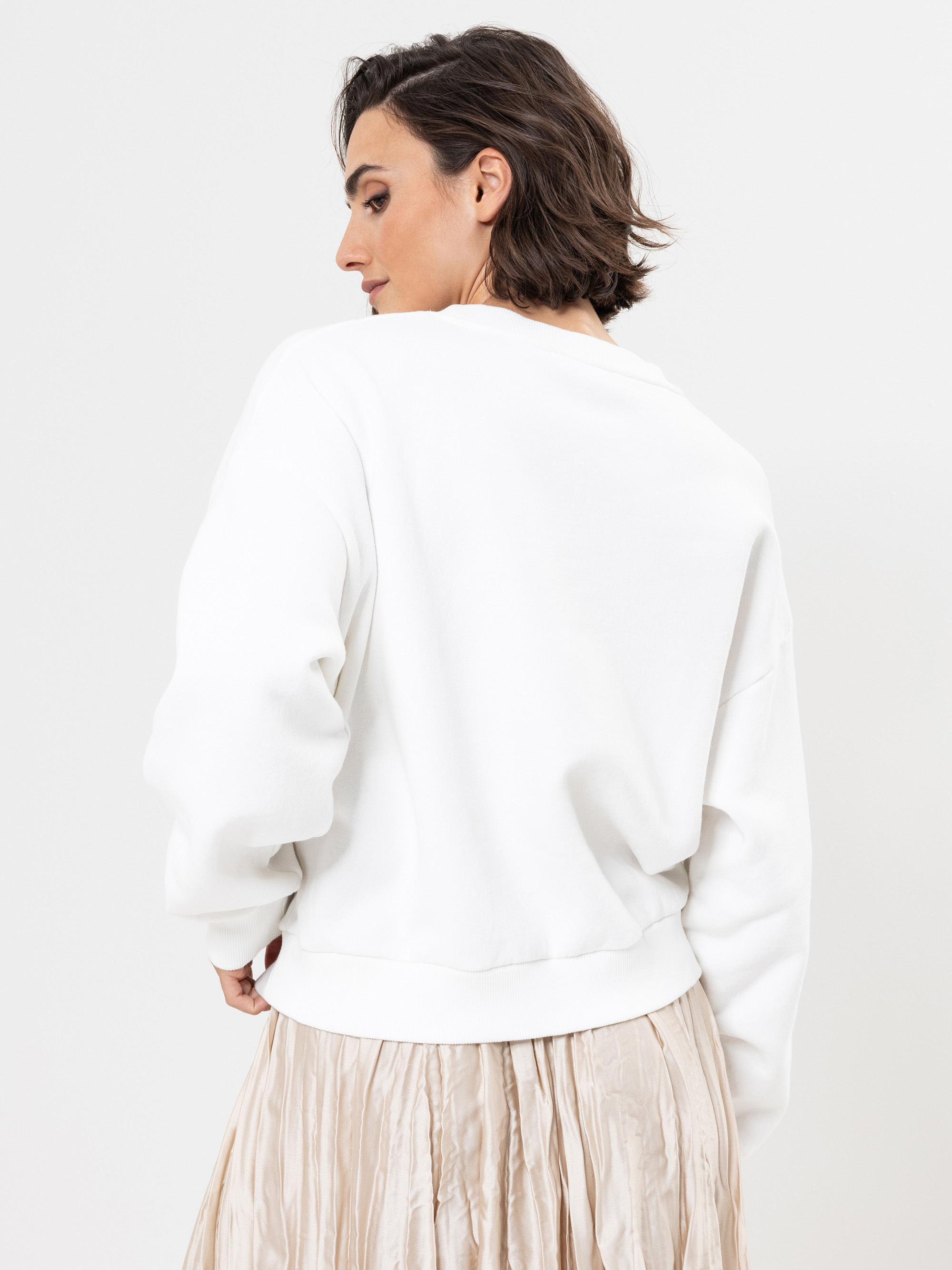 Eine Frau mit kurzen braunen Haaren trägt ein weißes Sweatshirt und einen beigen Faltenrock. Sie steht vor einem schlichten weißen Hintergrund und schaut über ihre Schulter.