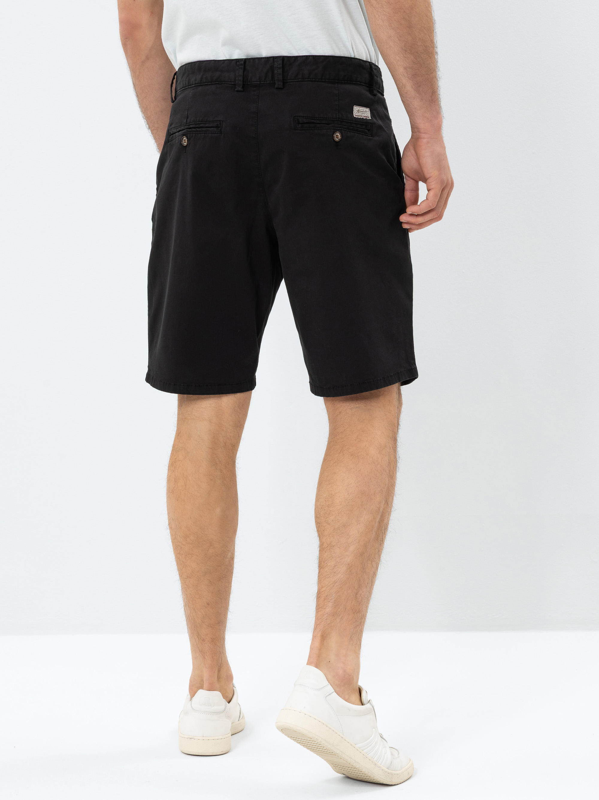 Ein Mann steht mit dem Rücken zur Kamera, trägt schwarze Shorts, ein helles Hemd und weiße Turnschuhe vor einem schlichten weißen Hintergrund.